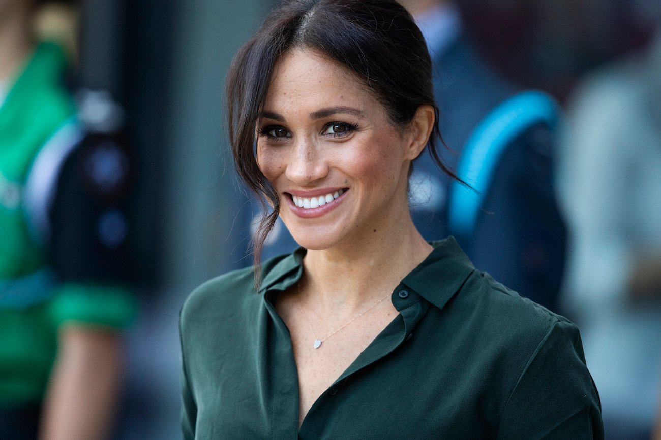 Meghan Markle smiling while wearing a dark green outfit and having an updo