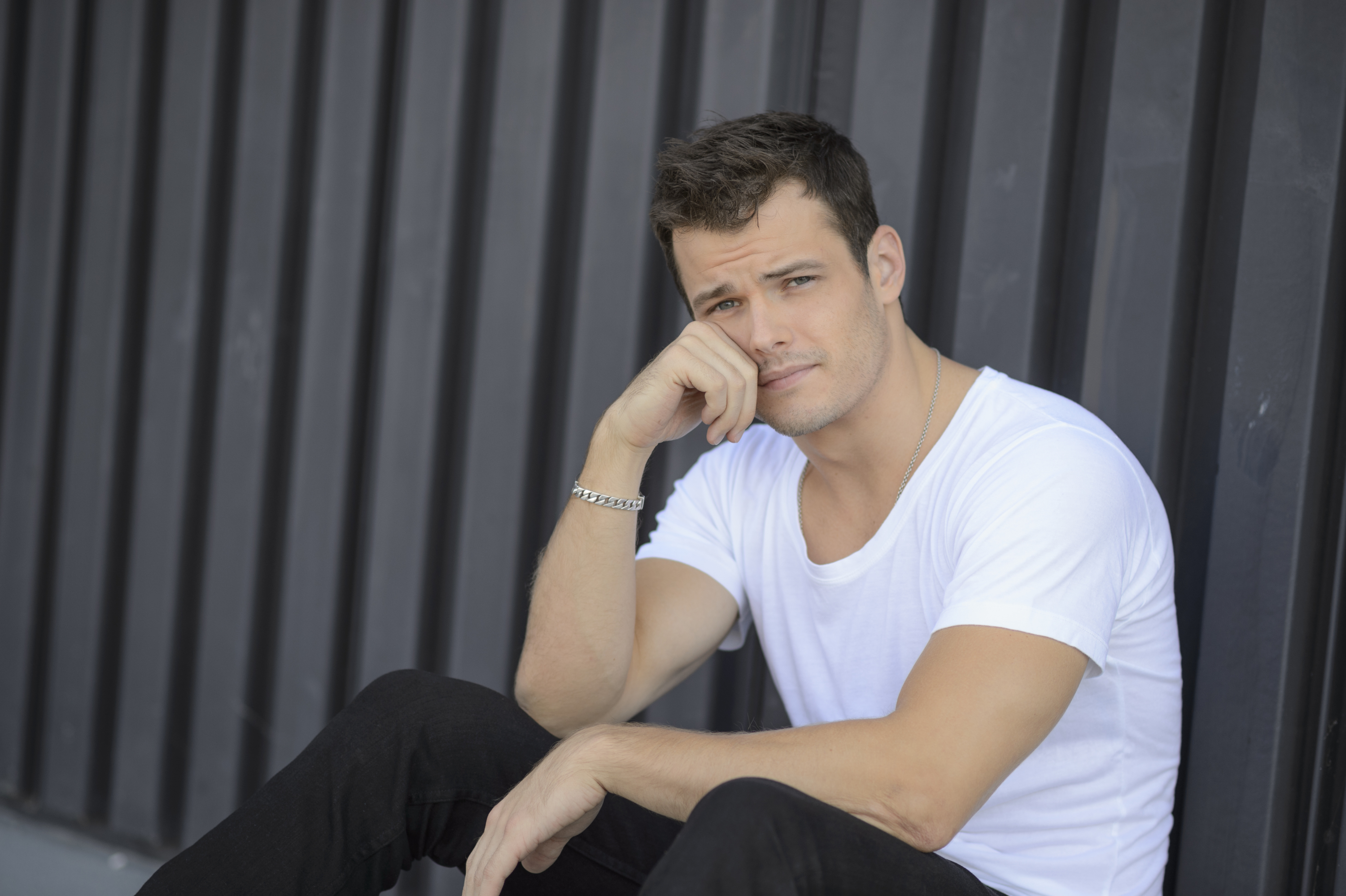 'The Young and the Restless' actor Michael Mealor wearing a white shirt and black pants; sits against a building.
