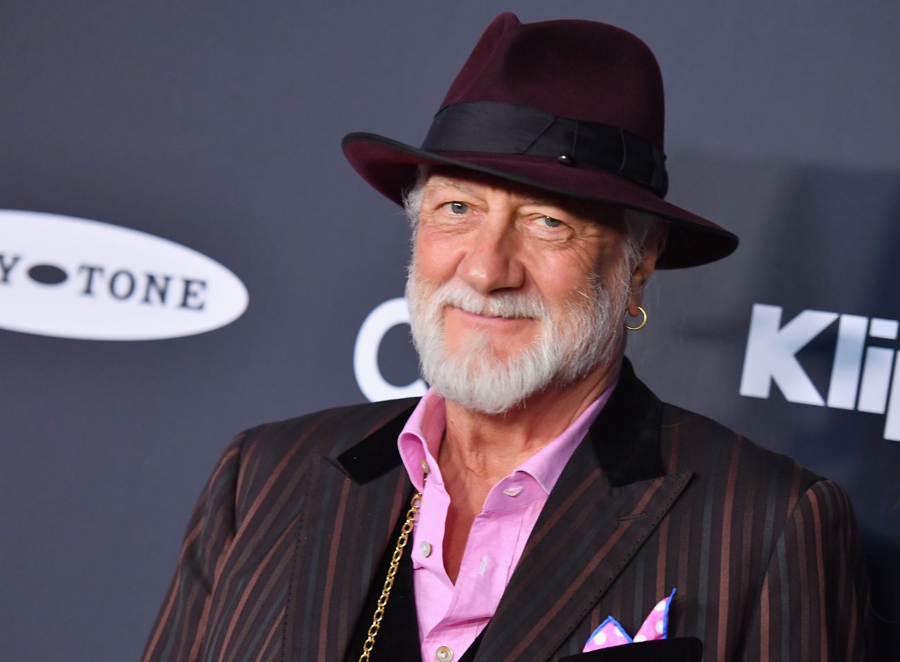 Mick Fleetwood wears a striped suit, gold jewelry, and a hat. Harry Styles made him the face of his beauty brand.