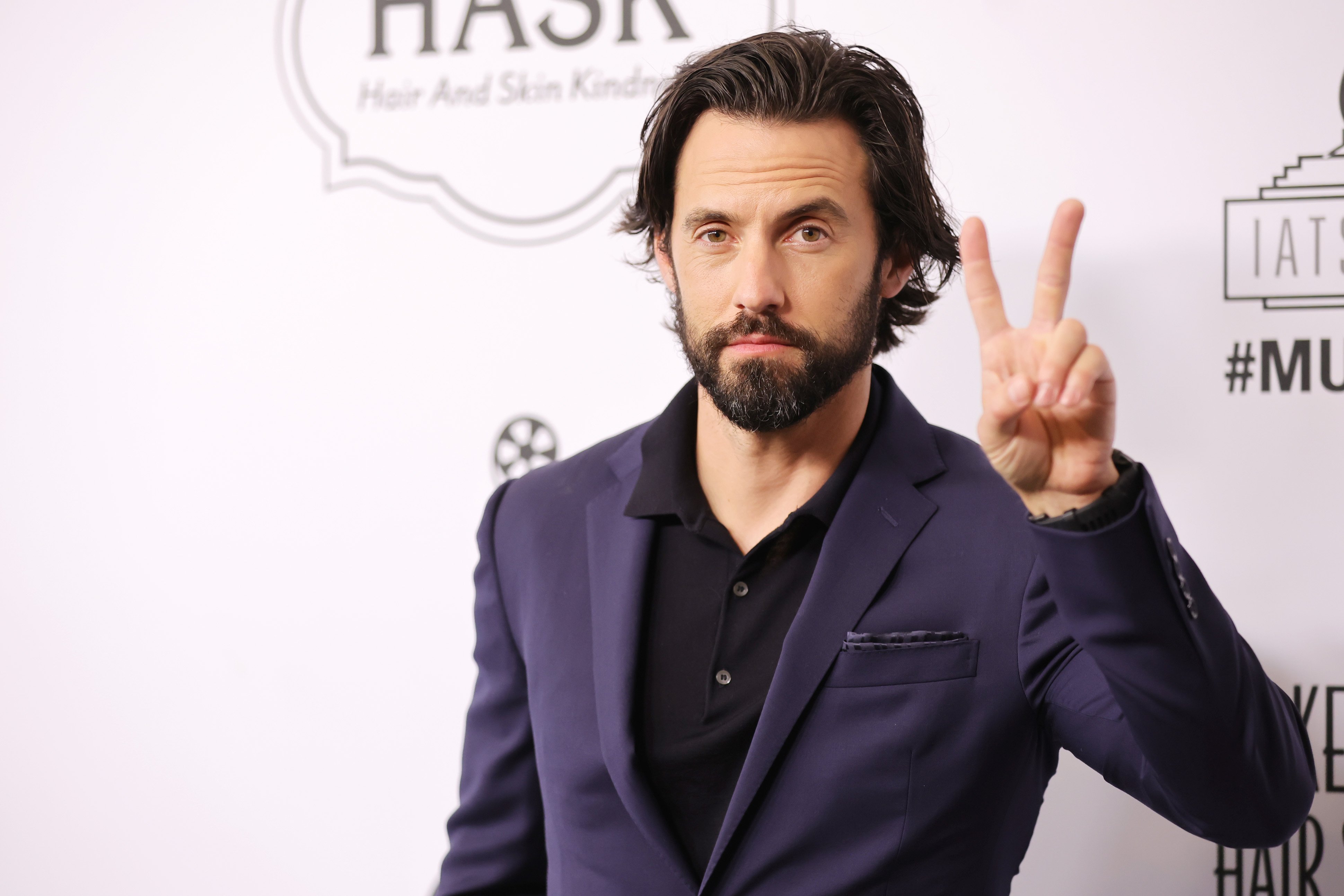 Milo Ventimiglia, star of 'This Is Us,' wears a dark blue suit over a black button-up shirt while holding up a peace sign.