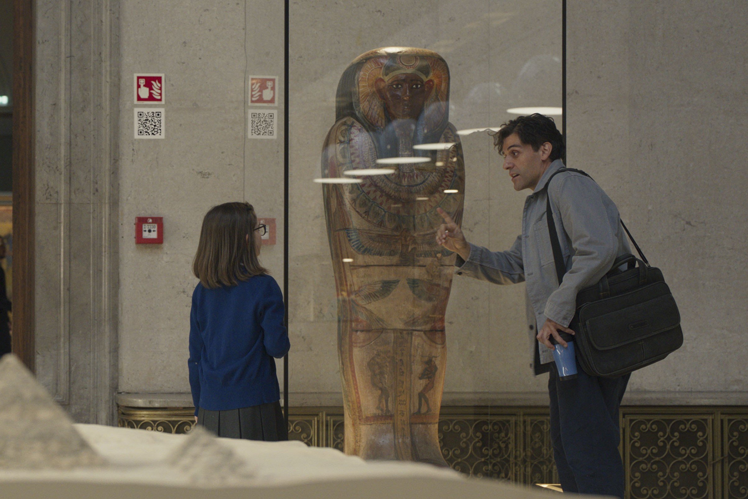 Oscar Isaac, in character as Steven Grant, shares a scene with a young girl in 'Moon Knight' Episode 1, which features an Easter egg. They stand in front of a sarcophagus. The girl wears a blue sweater and a gray skirt. Steven wears a gray jacket and blue pants, and has a black bag slung over his shoulder.