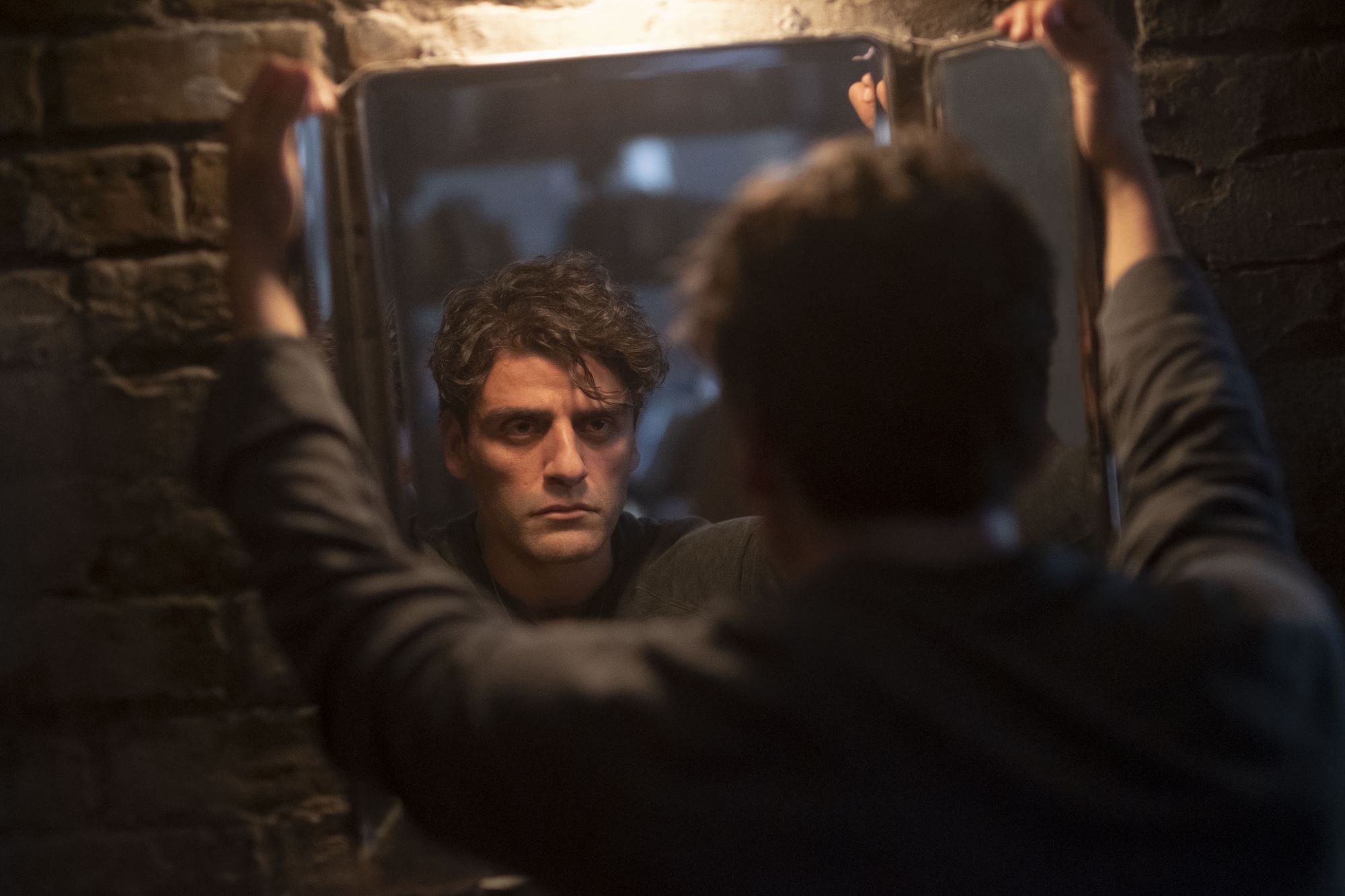 MCU's 'Moon Knight' star Oscar Isaac, in character as Steven Grant, stares at himself in the mirror. He wears a black long-sleeved shirt.