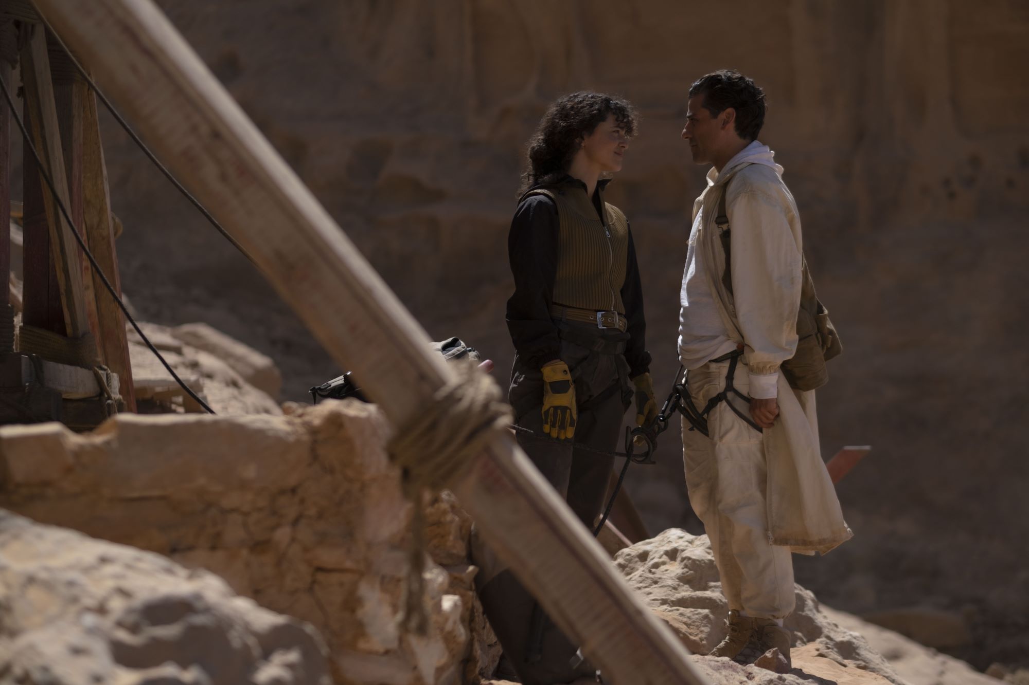 'Moon Knight' stars May Calamawy and Oscar Isaac, in character as Layla El-Faouly and Marc Spector, share a scene in a rocky terrain. Layla wears a brown vest over a black jacket and black pants. Marc wears a long white jacket over a white hoodie and white pants.