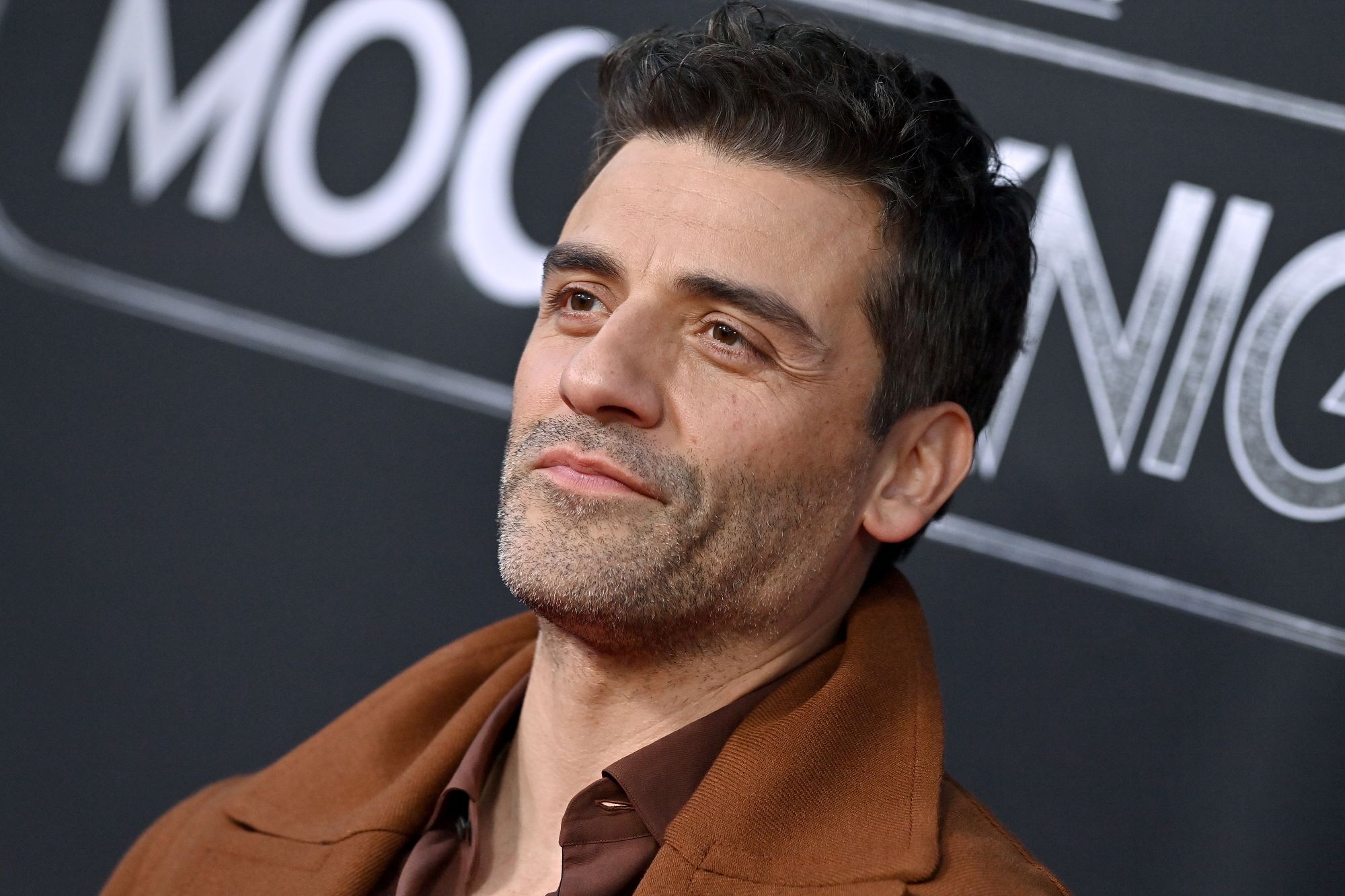 Oscar Isaac, star of 'Moon Knight,' wears a light brown coat over a dark brown button-up shirt.