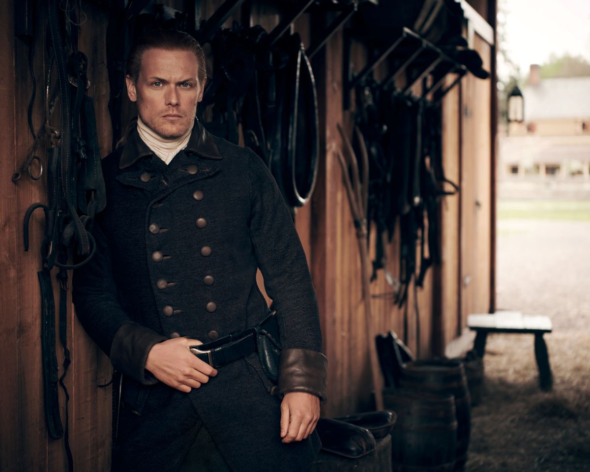 Sam Heughan in character as Jamie Fraser in a jacket and belt posing in a barn on Outlander