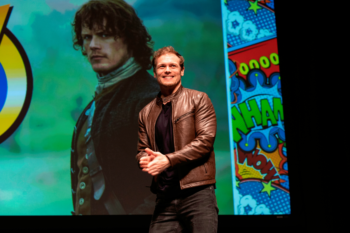Outlander Sam Heughan shadowed by a large image of Jamie Fraser speaks on stage during Wizard World Comic Con at Ernest N. Morial Convention Center on January 04, 2020