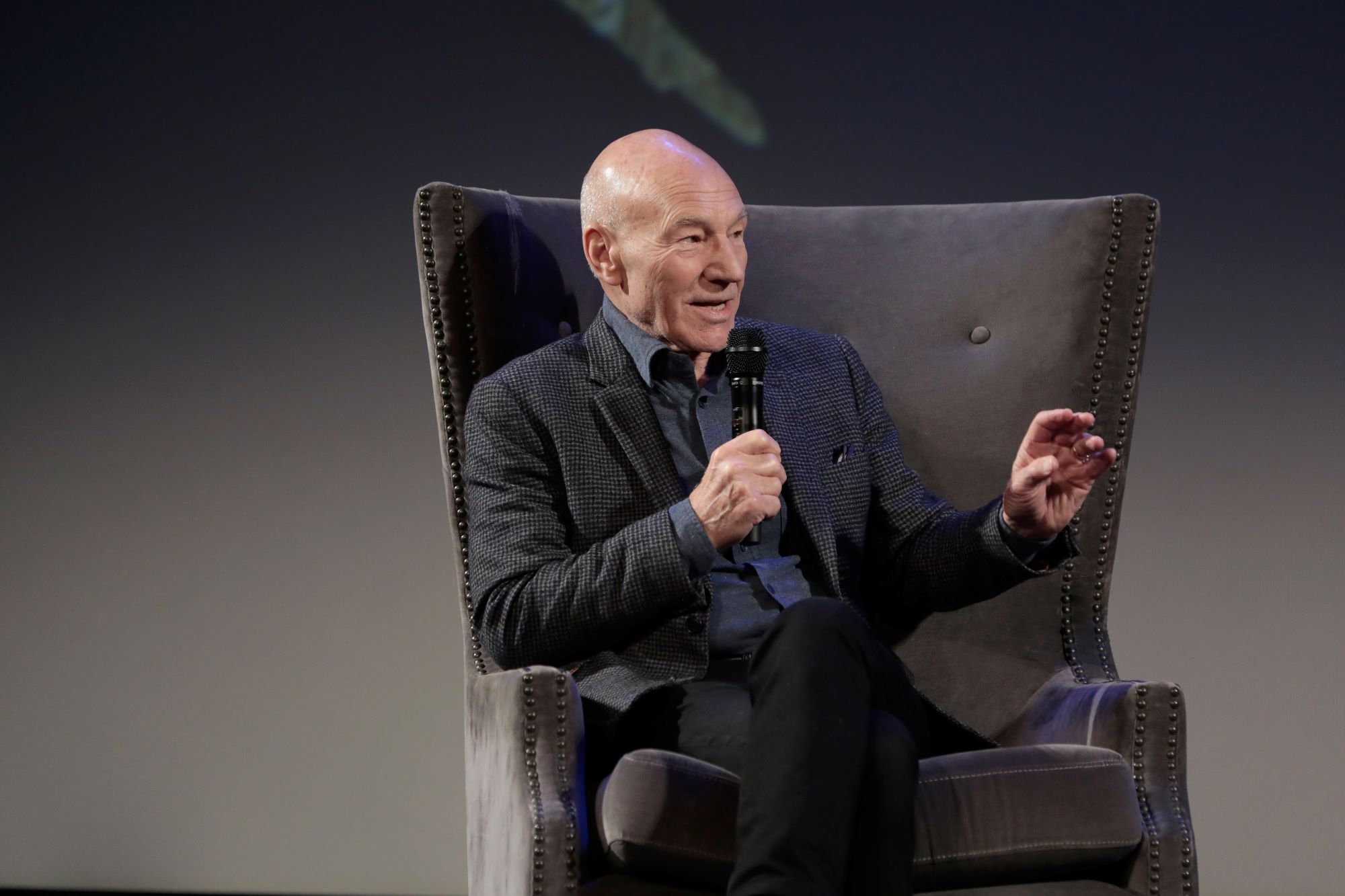Patrick Stewart, who stars in 'Doctor Strange 2,' wears a black jacket over a dark blue button-up shirt and black pants while he sits in a large chair and speaks into a microphone.
