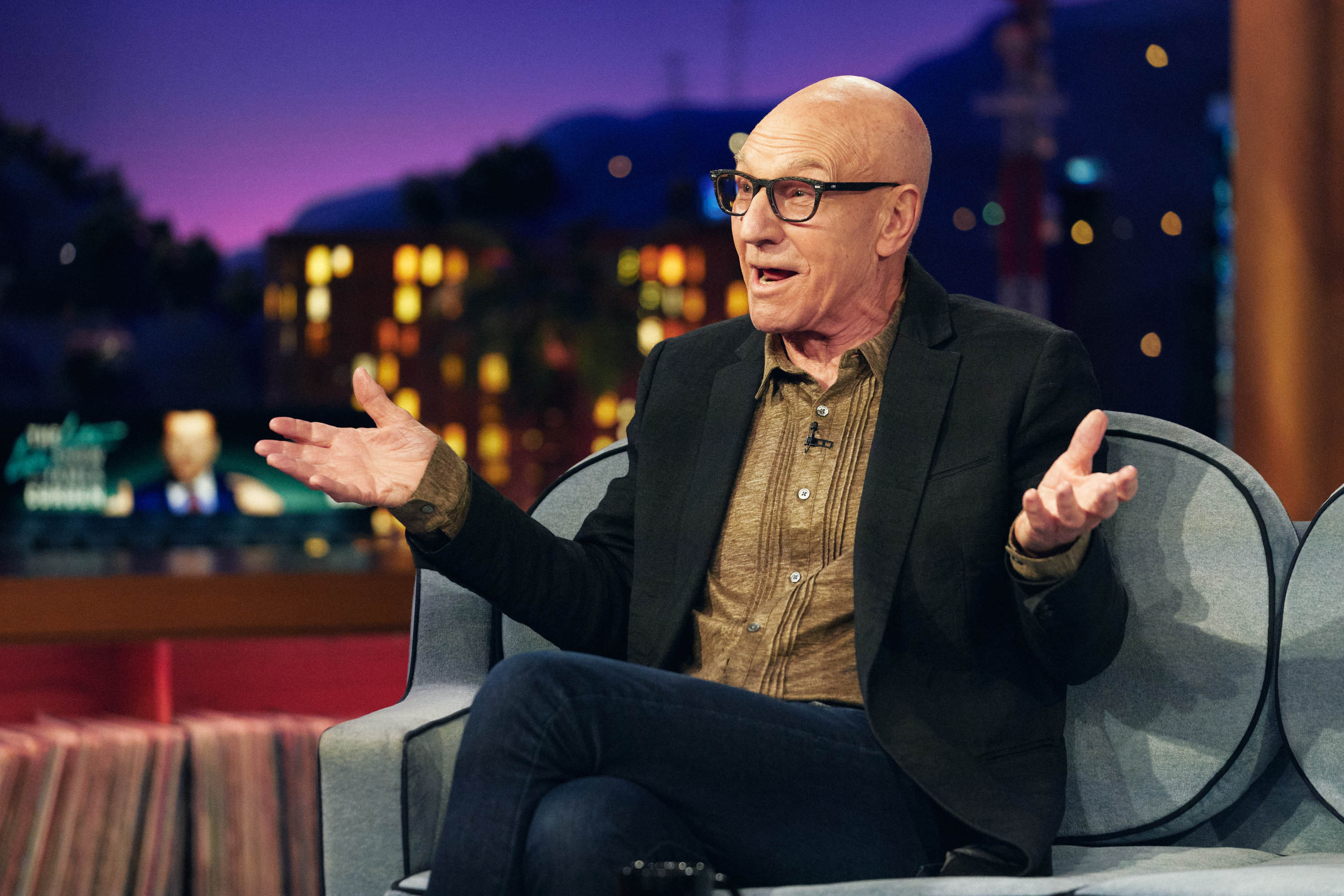 Patrick Stewart, who many fans believe is in 'Doctor Strange 2,' wears a black suit jacket over a light brown button-up shirt and jeans. He is also wearing black-framed glasses.