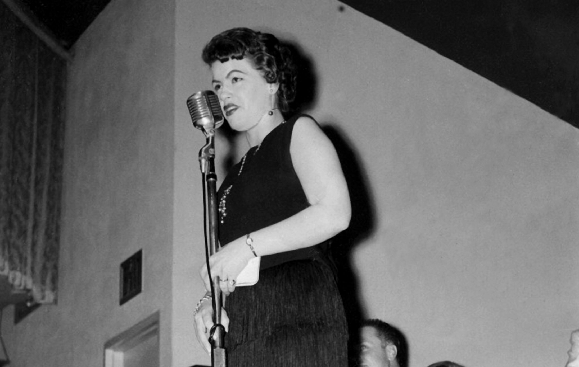 Patsy Cline in a sleeveless dress singing into a microphone c. 1950