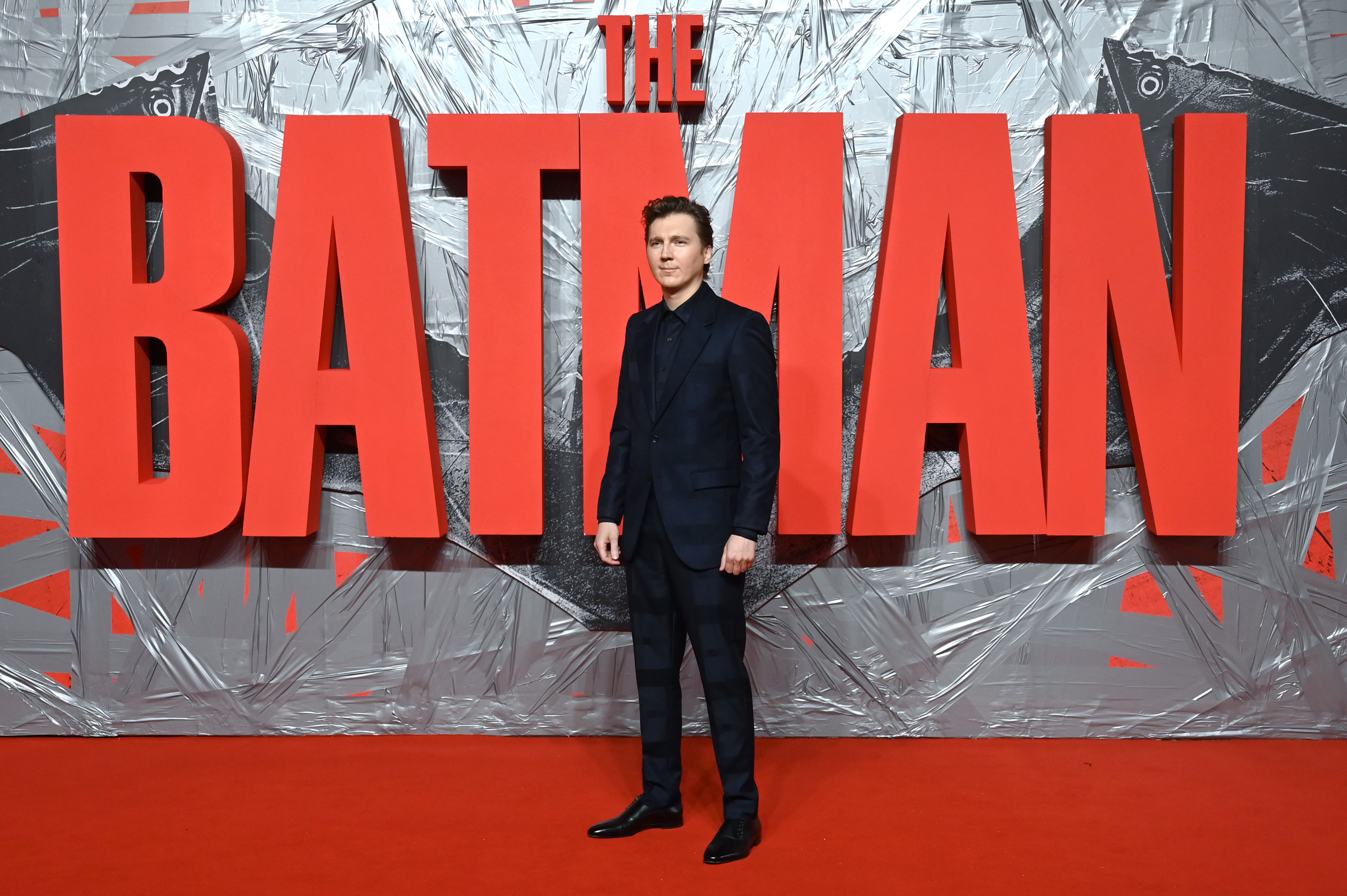 Paul Dano, who plays the Riddler in 'The Batman,' wears a black suit while standing in front of a sign for 'The Batman.'