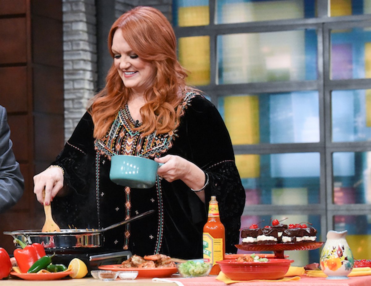 Pioneer Woman Ree Drummond smiles as she holds a pot in one hand and stirs another with a wooden spoon