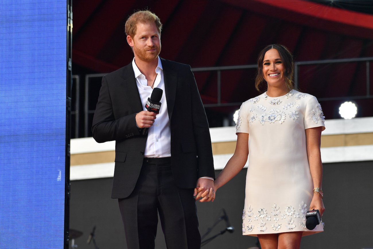 Prince Harry wearing a suit and holding hands with Meghan Markle, who is wearing an off-white dress