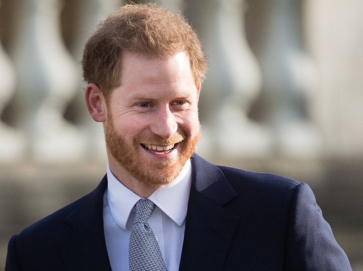 Prince Harry smiles and looks on wearing a navy blazer and blue tie