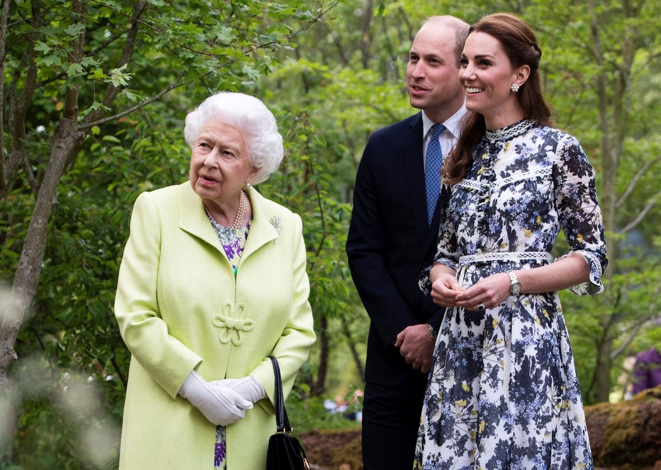 Queen Elizabeth standing with Prince William and Kate Middleton, all looking toward the left side of the picture
