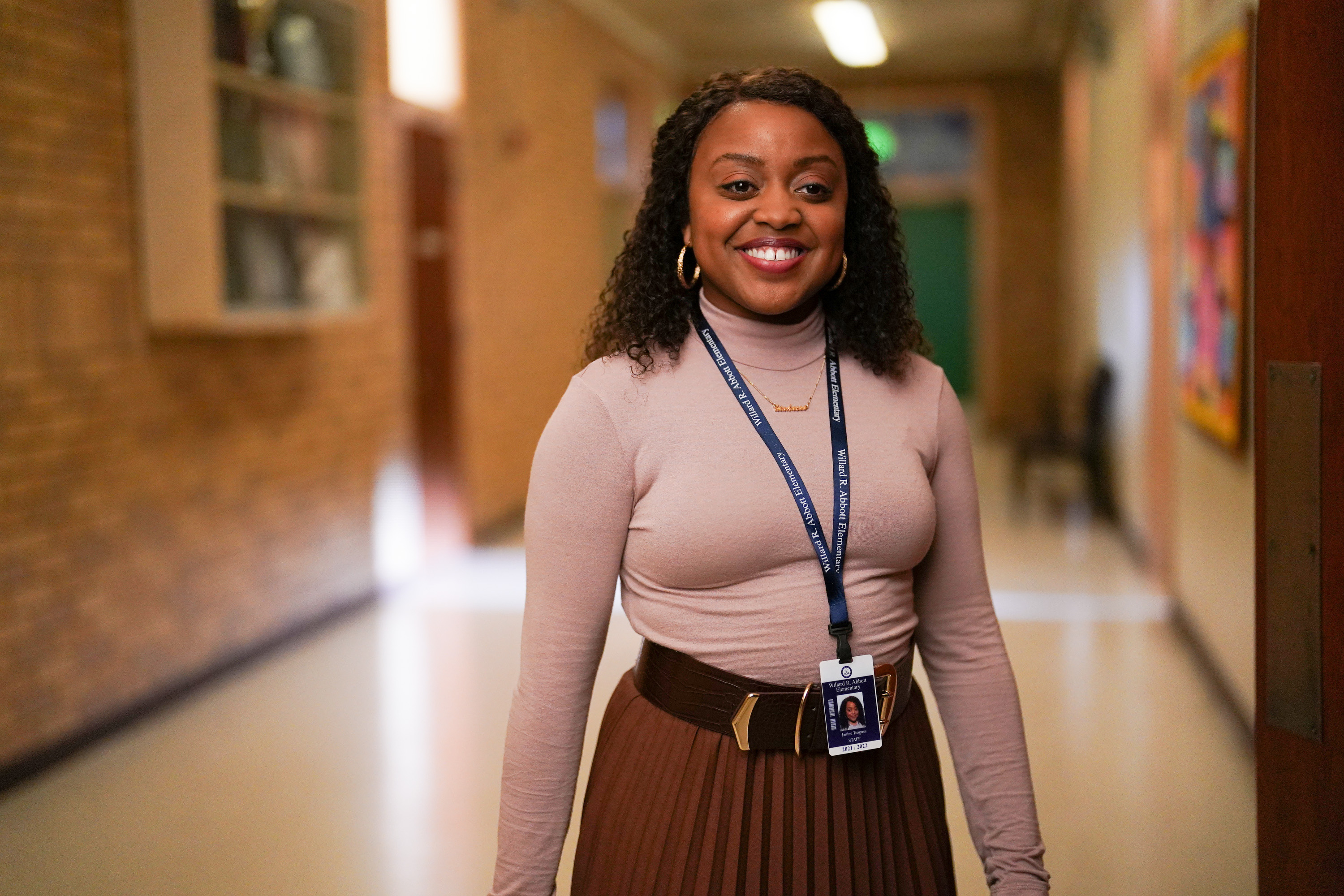 'Abbott Elementary' star Quinta Brunson, in character as Janine Teagues, wears a light pink turtleneck, a brown skirt, and a lanyard with her ID.