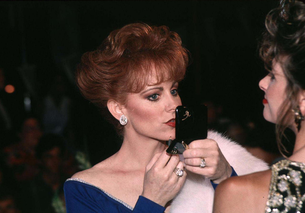 Reba McEntire in an off-the-shoulder dress, looking into a mirror and putting on lipstick
