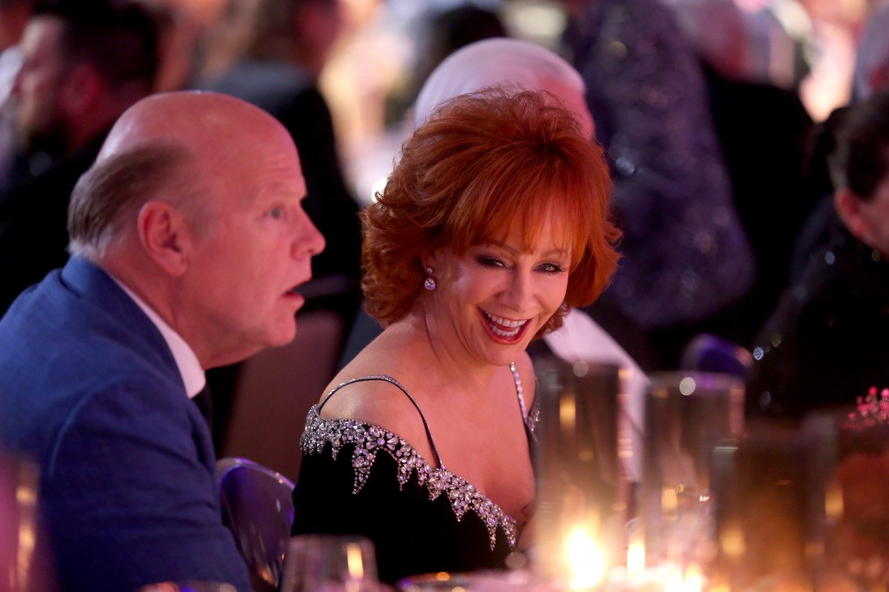 Rex Linn and Reba McEntire seated at a table as McEntire looks to the camera and smiles 