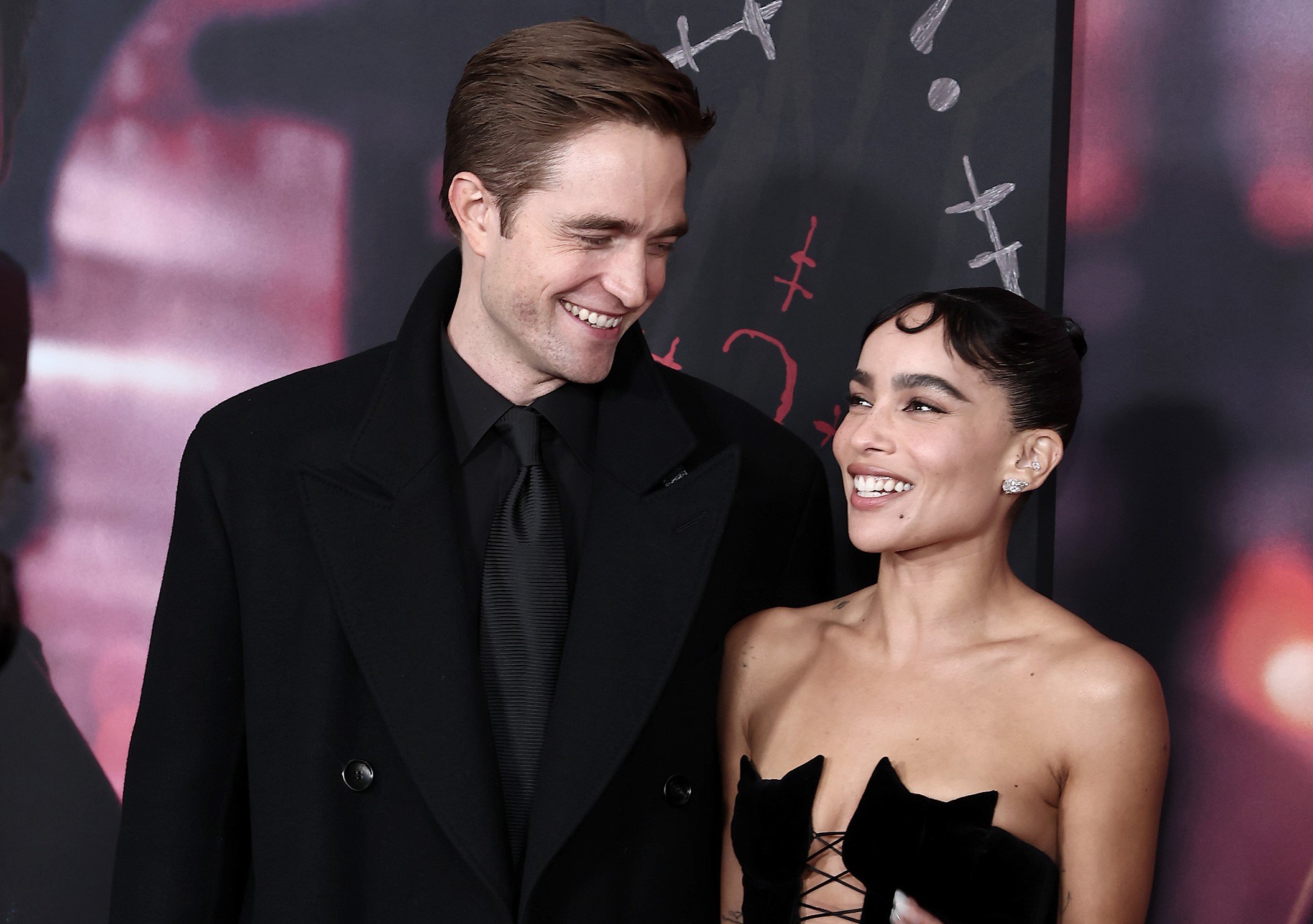 'The Batman' stars Robert Pattinson and Zoë Kravitz pose for photos together on the red carpet of the premiere. Pattinson wears a black coat over a black button-up shirt and black tie. Kravitz wears a strapless black dress with a neckline shaped like two cats.