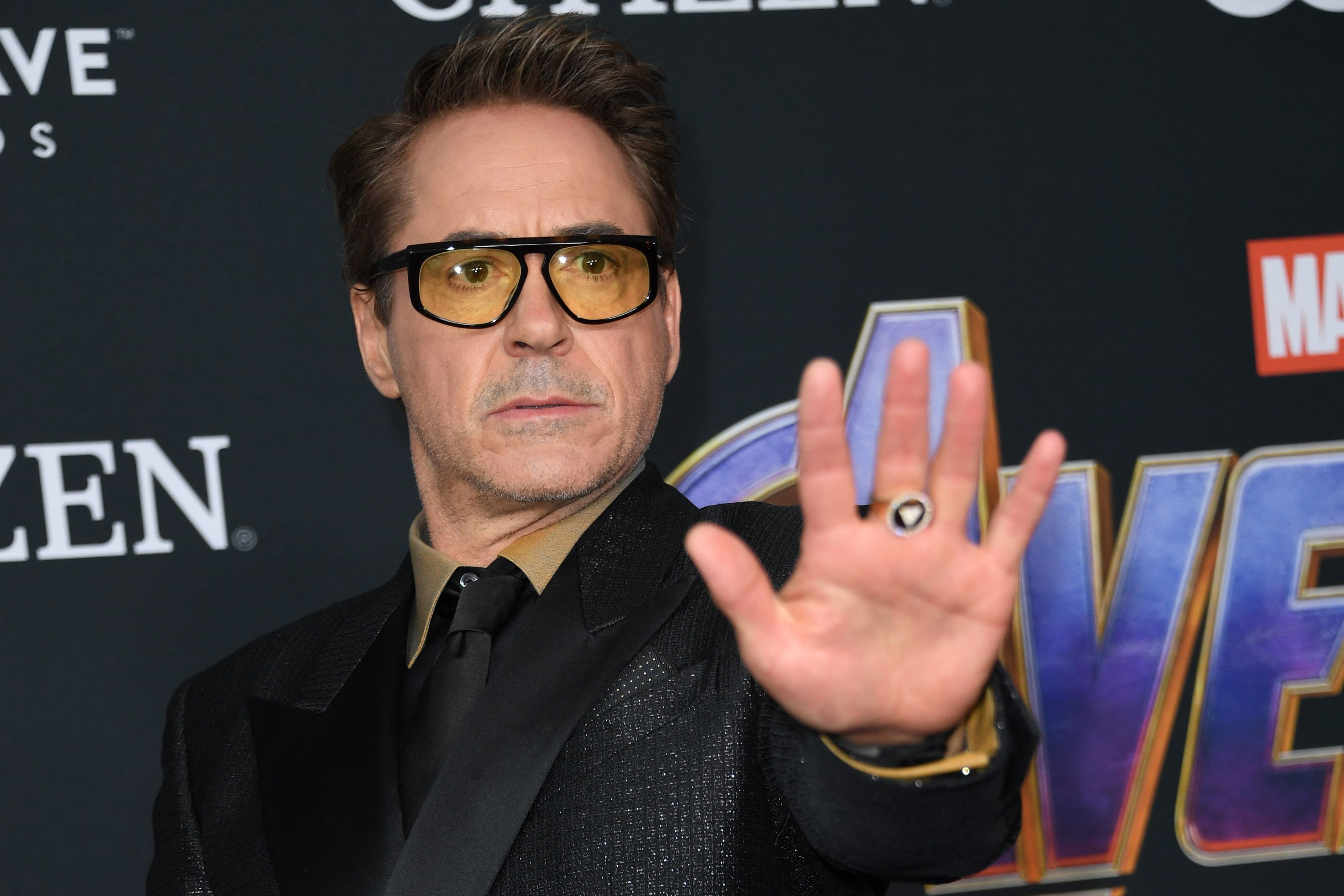 Robert Downey Jr., who played Iron Man in 'Avengers: Endgame,' holds his arm in front of him with his palm facing the camera. The actor wears a black suit over a gold and black button-up shirt and black tie, and yellow-tinted glasses.