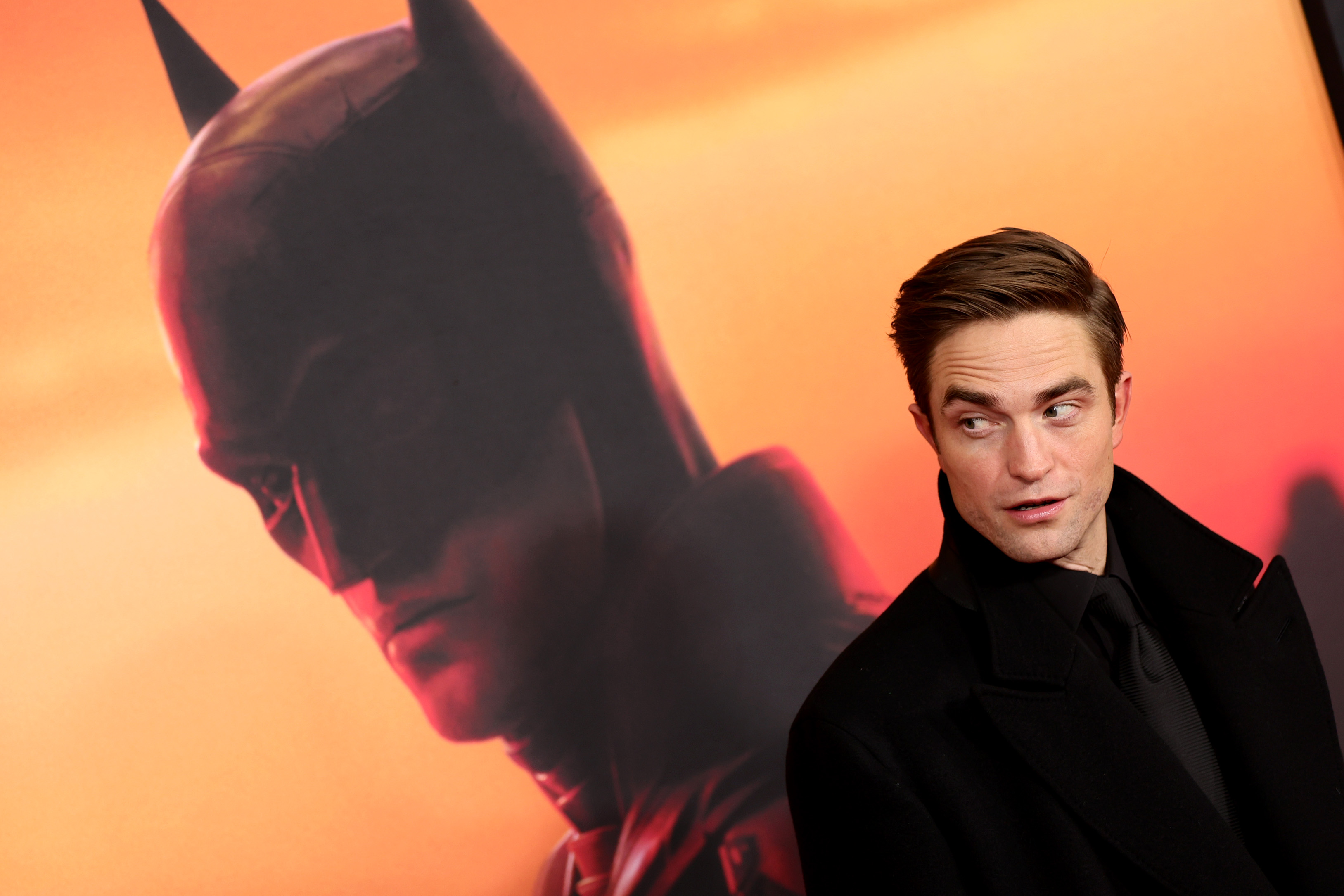 'The Batman' star Robert Pattinson stands in front of a poster for the film wearing a black coat over a black button-up shirt and black tie.