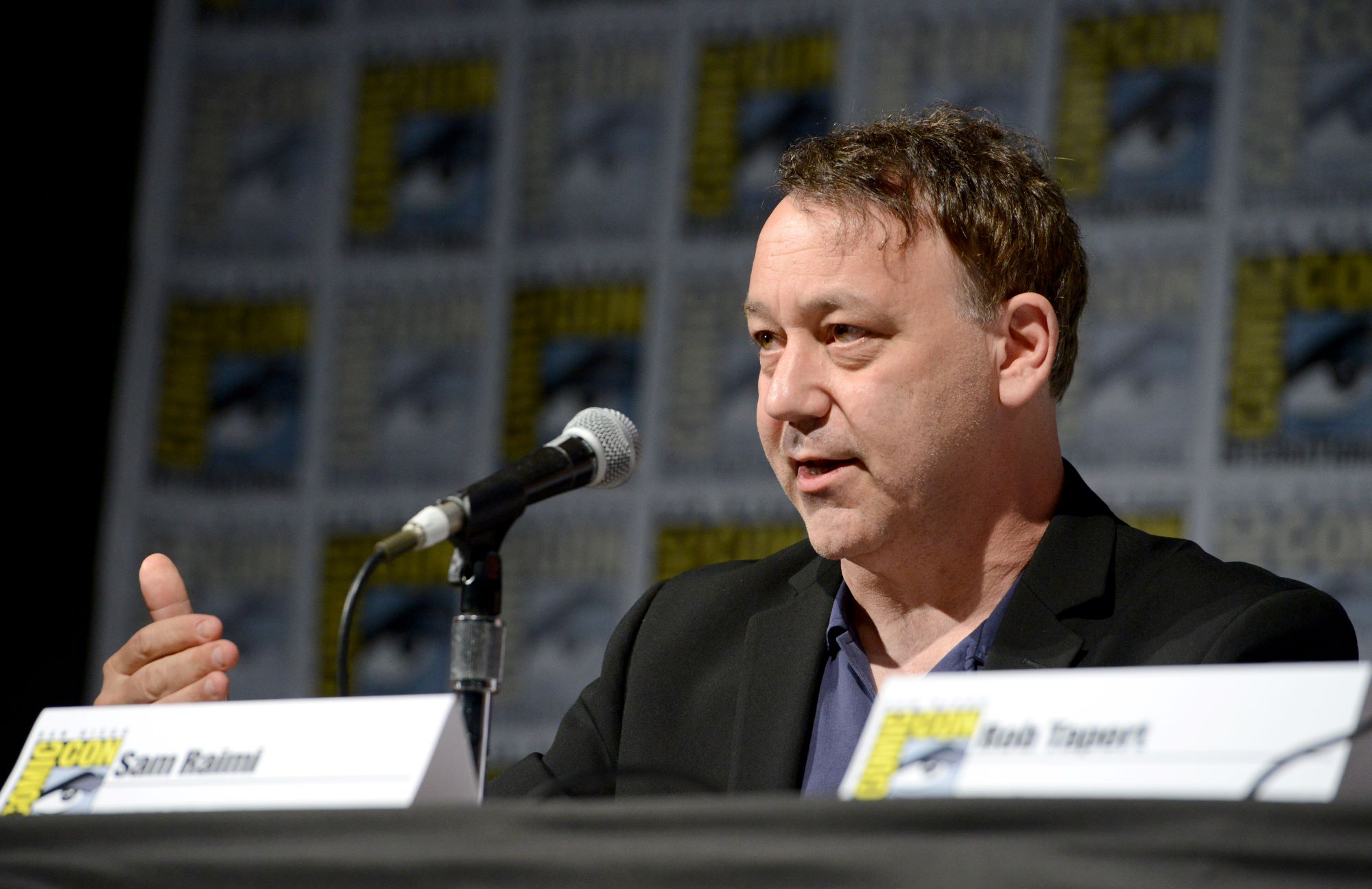 'Doctor Strange 2' director Sam Raimi wears a black suit over a blue button-up shirt while speaking onstage at a Comic Con panel.