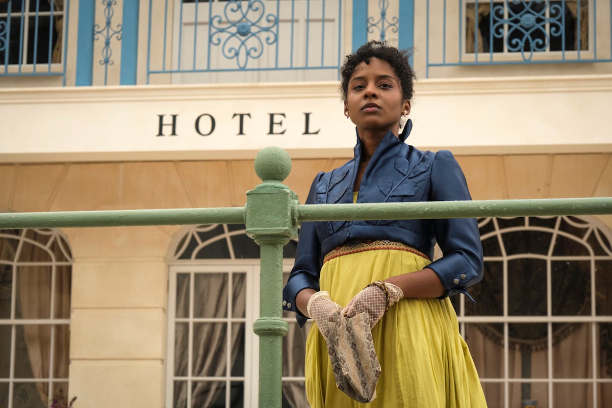 Georgiana,,wearing a blue jacket and yellow skirt, standing in front of a hotel in 'Sanditon' Season 2