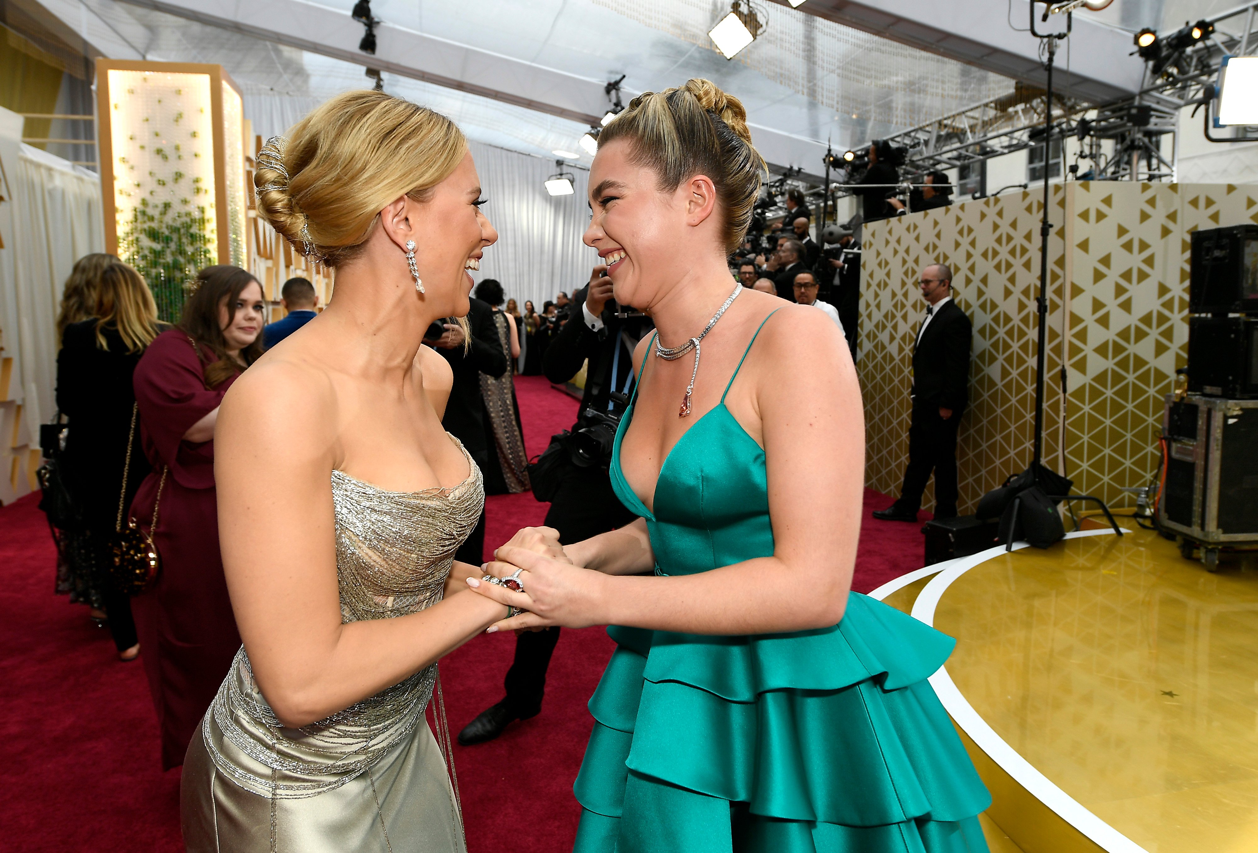 'Black Widow' stars Scarlett Johansson and Florence Pugh smile at one another and hold hands on the red carpet. Johansson wears a strapless sparkly silver dress. Pugh wears a teal dress.