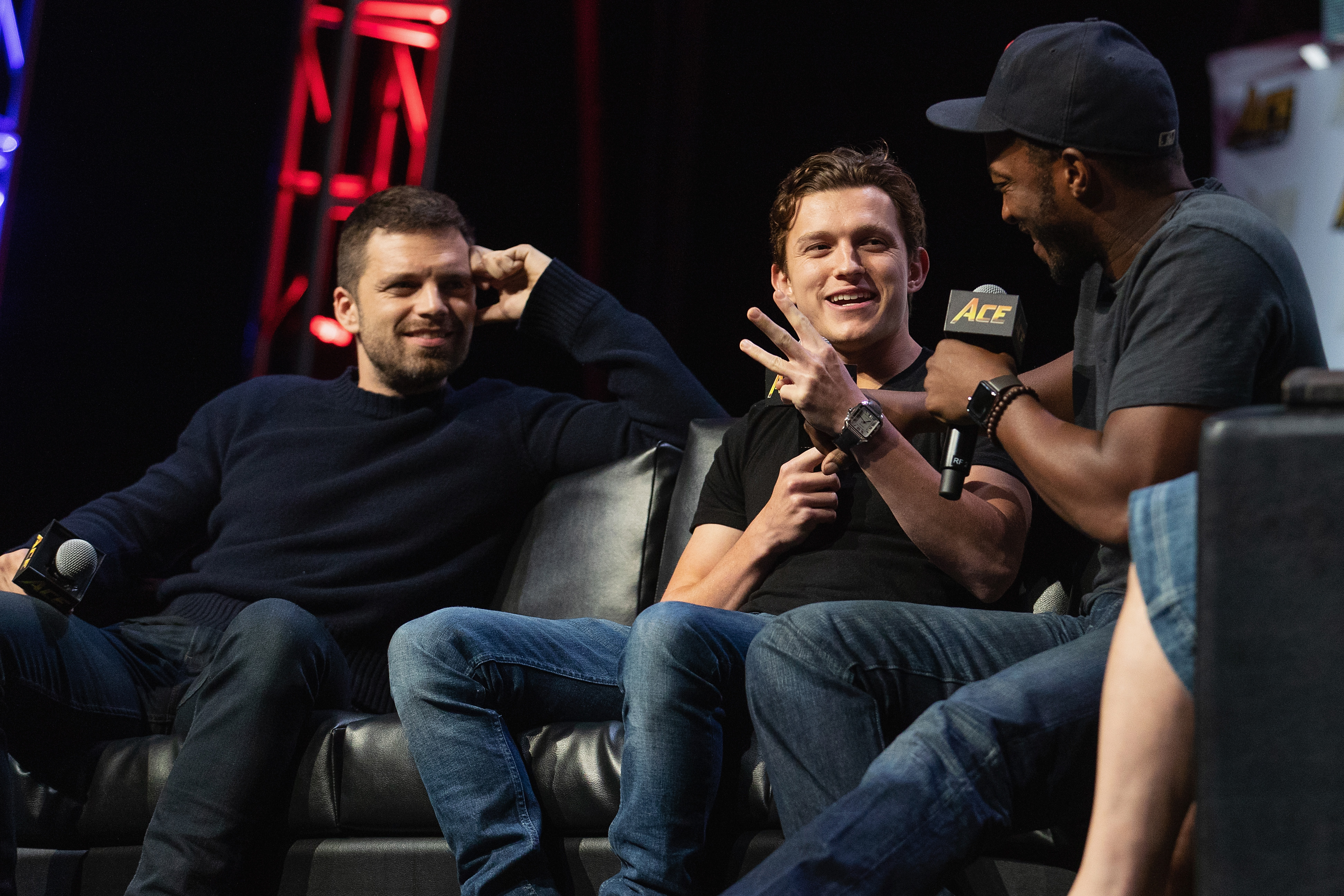 'The Falcon and the Winter Soldier' stars Sebastian Stan and Anthony Mackie speak onstage during a convention with Tom Holland. Stan wears a dark blue sweater and jeans. Holland wears a black shirt and jeans. Mackie wears a dark gray shirt, jeans, and a black baseball cap.