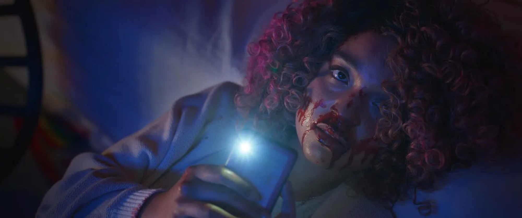 'Sissy' Aisha Dee as Cecilia using phone flashlight with blood on her face looking scared