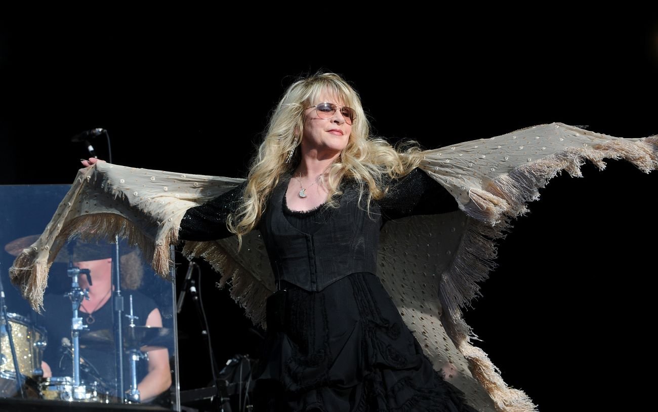Stevie Nicks wears sunglasses and spreads her arms. She wears a shawl.