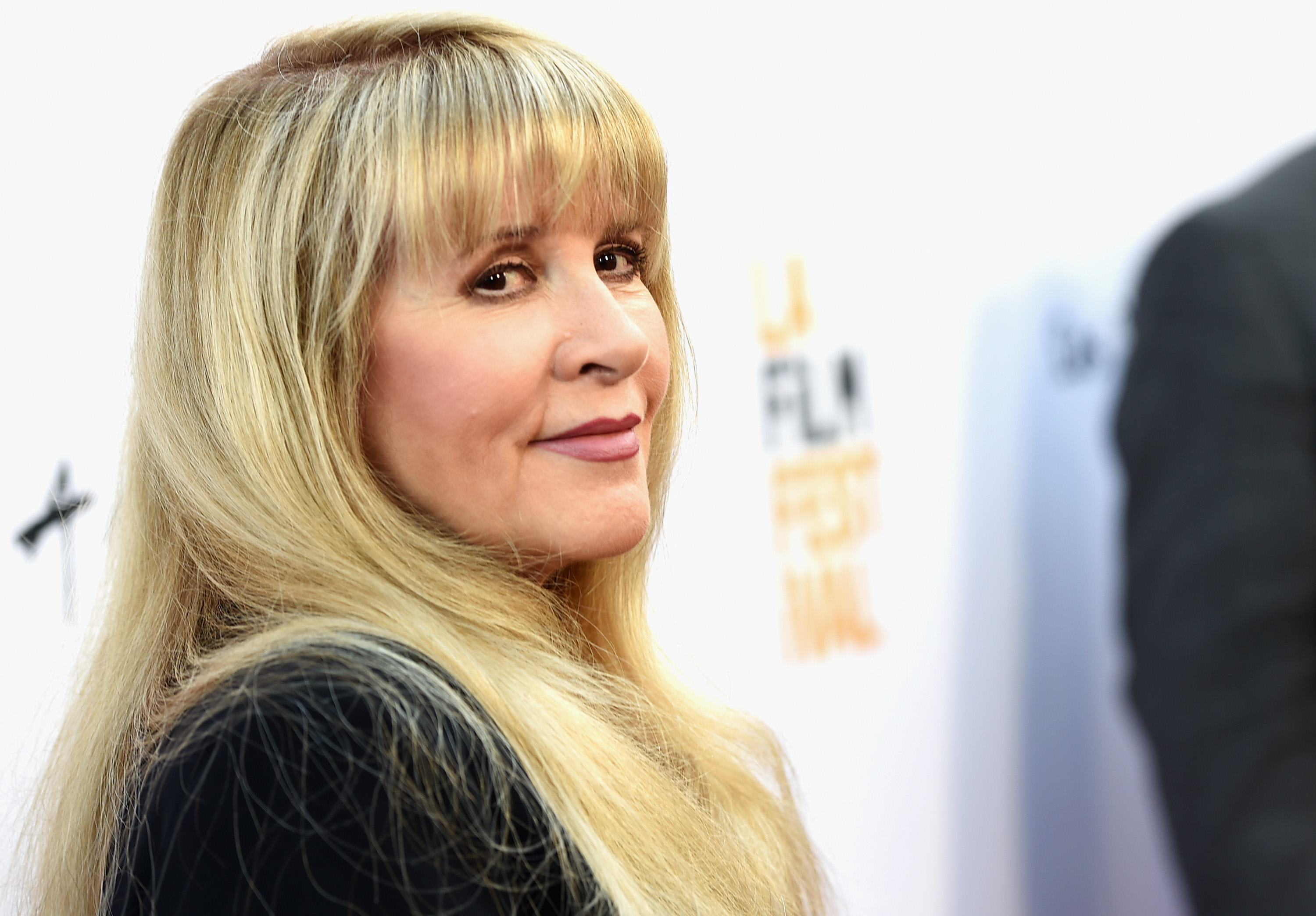 Stevie Nicks wears a black shirt and stands in front of a white background. 