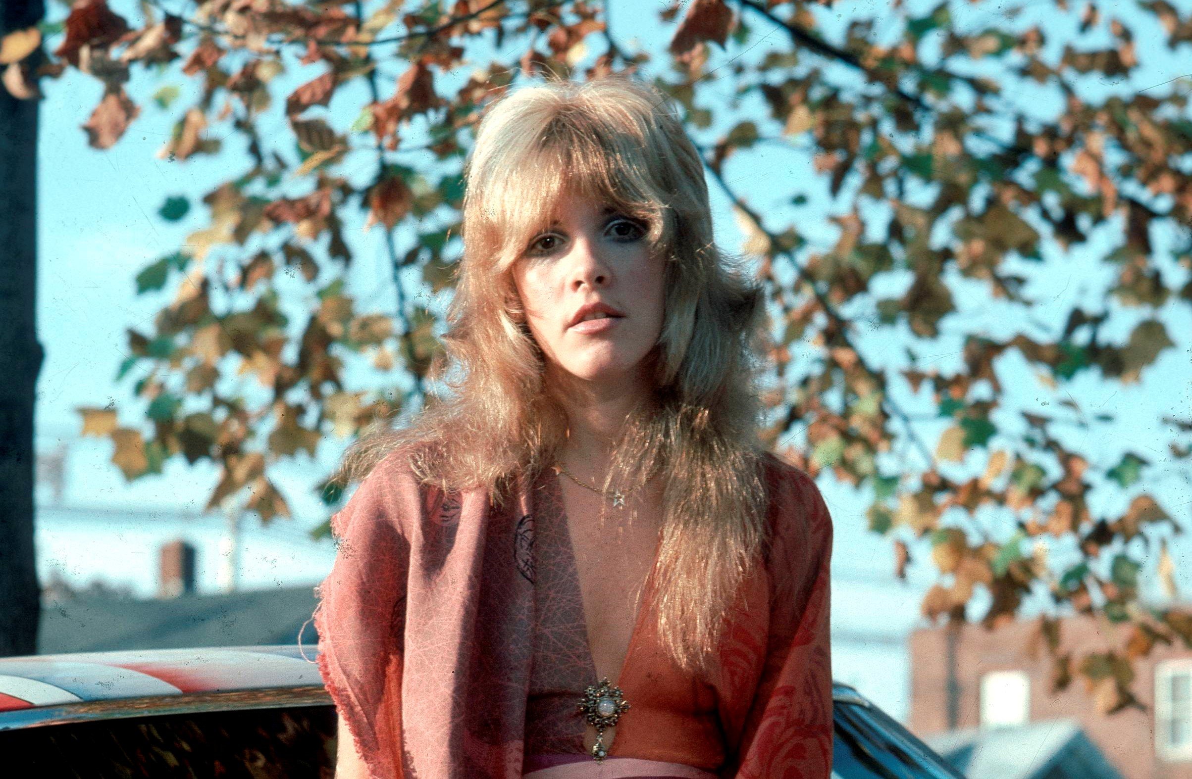 A young Stevie Nicks wears an orange shirt and sits on a car parked in front of a tree.