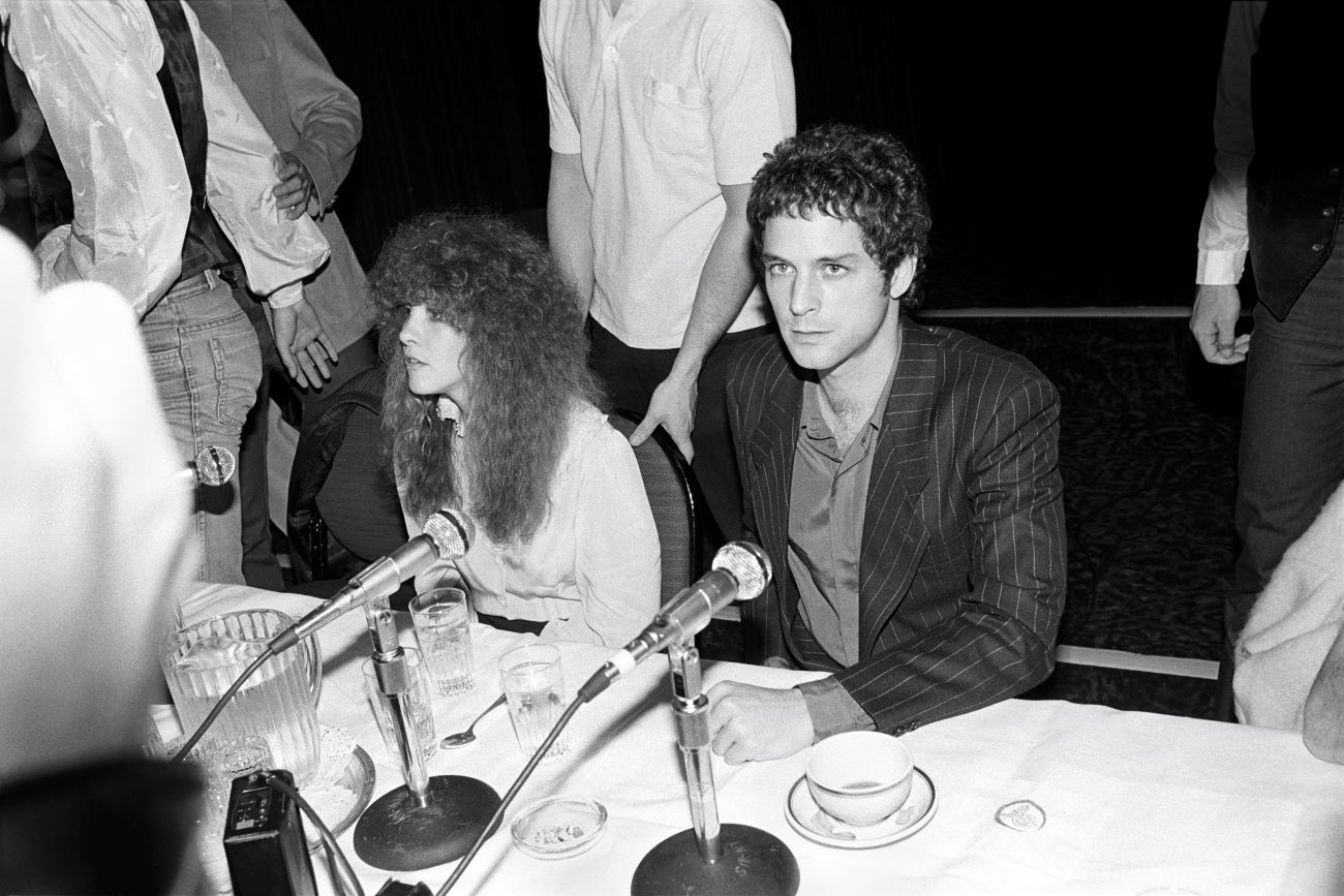 A black and white photo of Fleetwood Mac artists Stevie Nicks and Lindsey Buckingham sitting at a table in front of a microphones. 