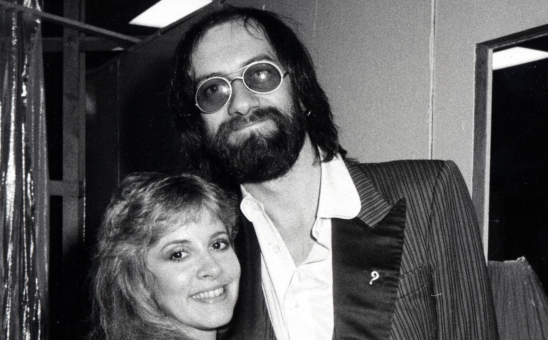 A black and white photo of Stevie Nicks and Mick Fleetwood smiling together. He wears sunglasses.