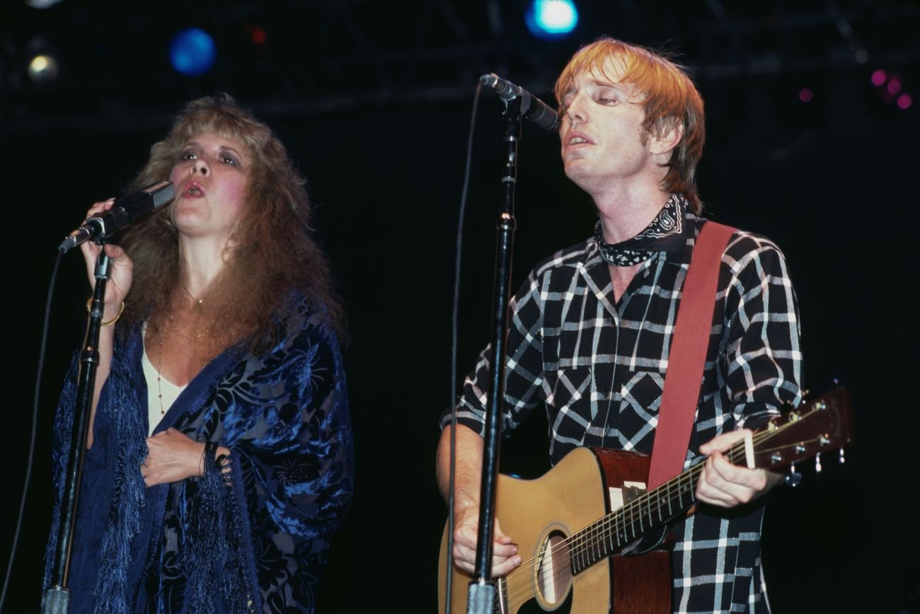 Stevie Nicks wears a blue shirt and sings into a microphone. Tom Petty wears a black plaid shirt and plays guitar while singing.