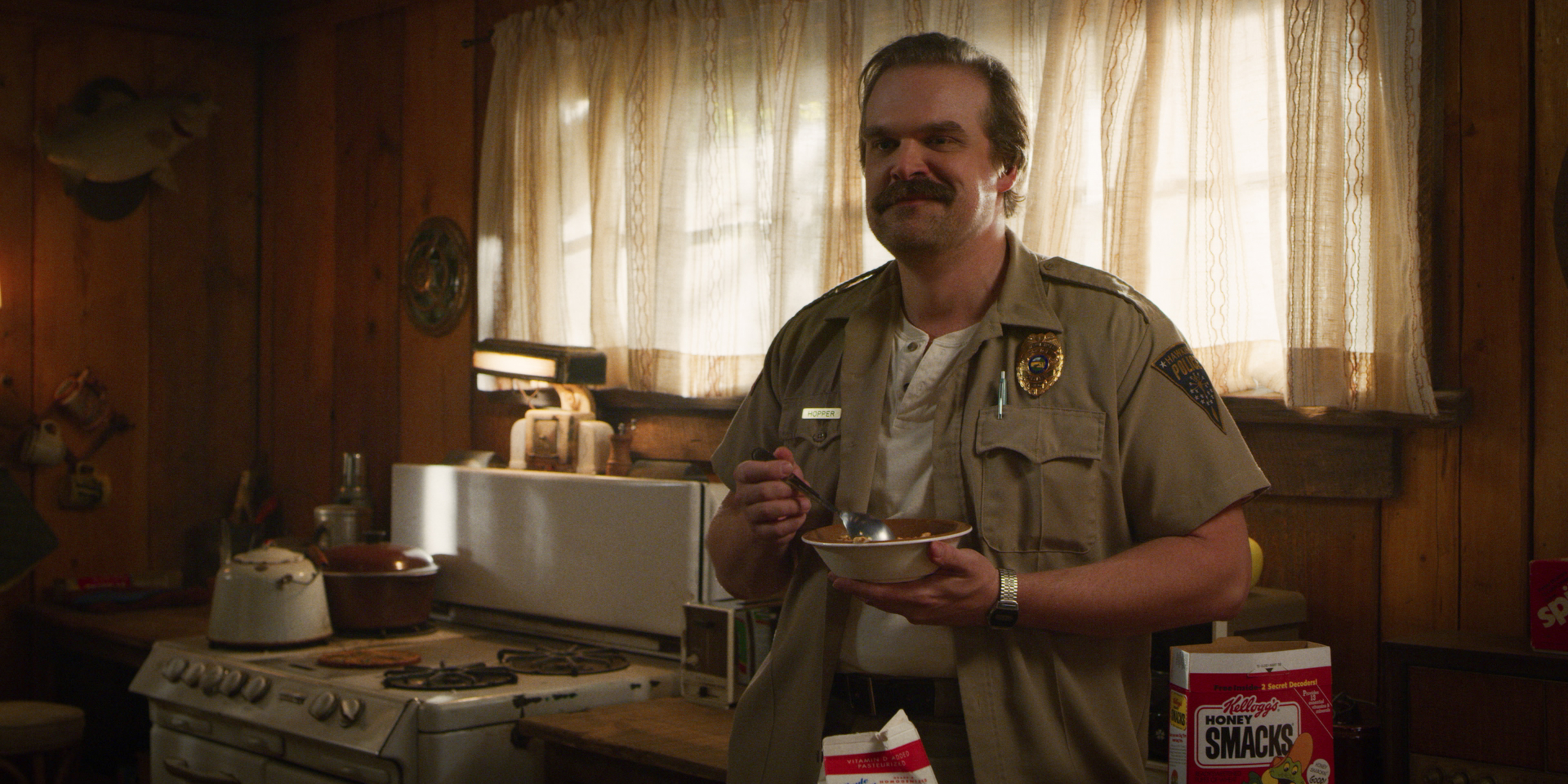 'Stranger Things' Season 3 production still of Chief Jim Hopper (David Harbour) leaning against a counter while eating a bowl of cereal.