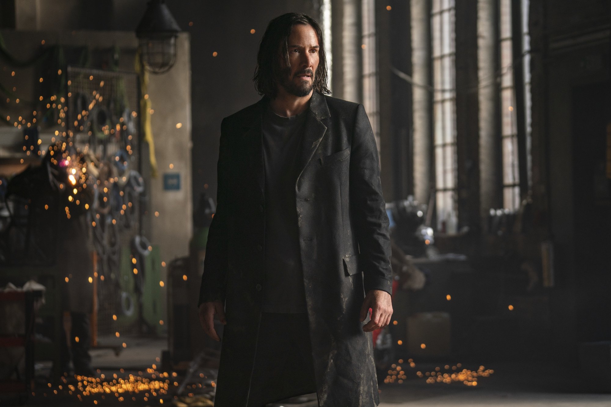 Stunt coordinator Scott Roger's 'The Matrix Resurrections' Keanu Reeves as Neo_Thomas A. Anderson standing wearing a trench coat with sparks flying in the background