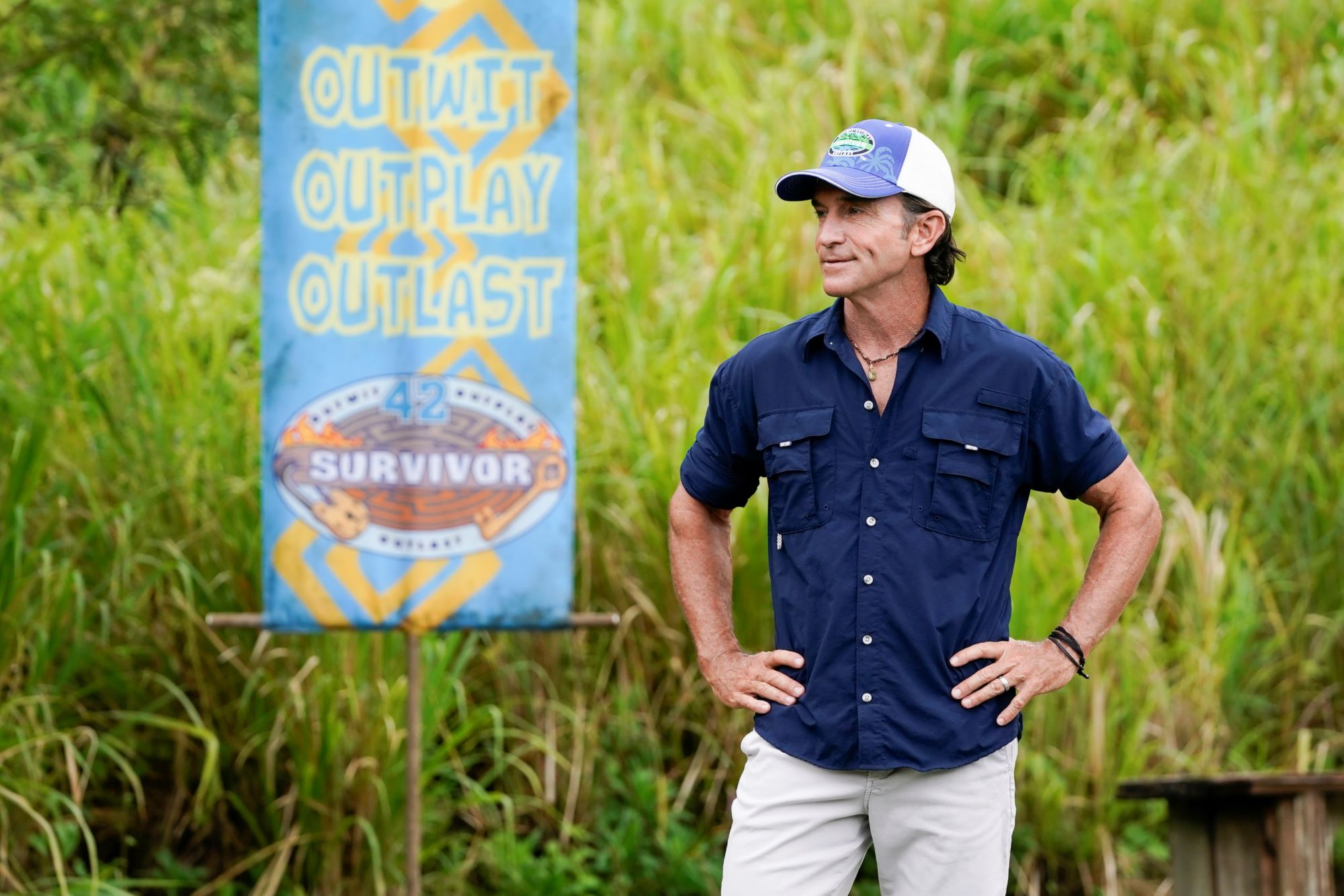 Jeff Probst, who hosts a new episode of 'Survivor' Season 42 tonight, March 23, wears a dark blue button-up shirt with the sleeves rolled up to his elbows, light beige pants, and a blue, green, and white 'Survivor' baseball hat.