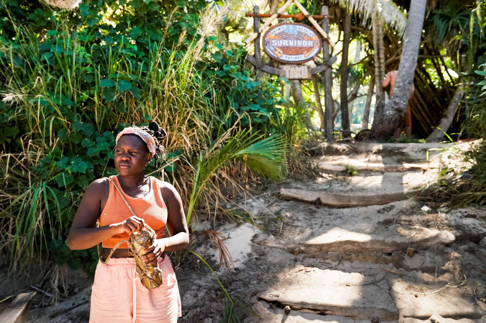 Maryanne Oketch, a castaway in 'Survivor' Season 42 who admitted to having a crush on Zach, wears an orange tank top, light pink shorts, and her orange buff wrapped around her head.