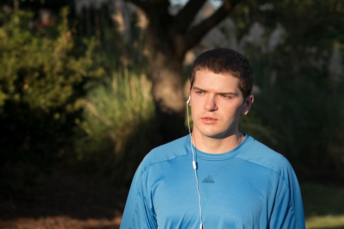 Colton Ryan as Conrad Roy III in The Girl from Plainville. Conrad wears a blue t-shirt and has a headphone in one ear. 