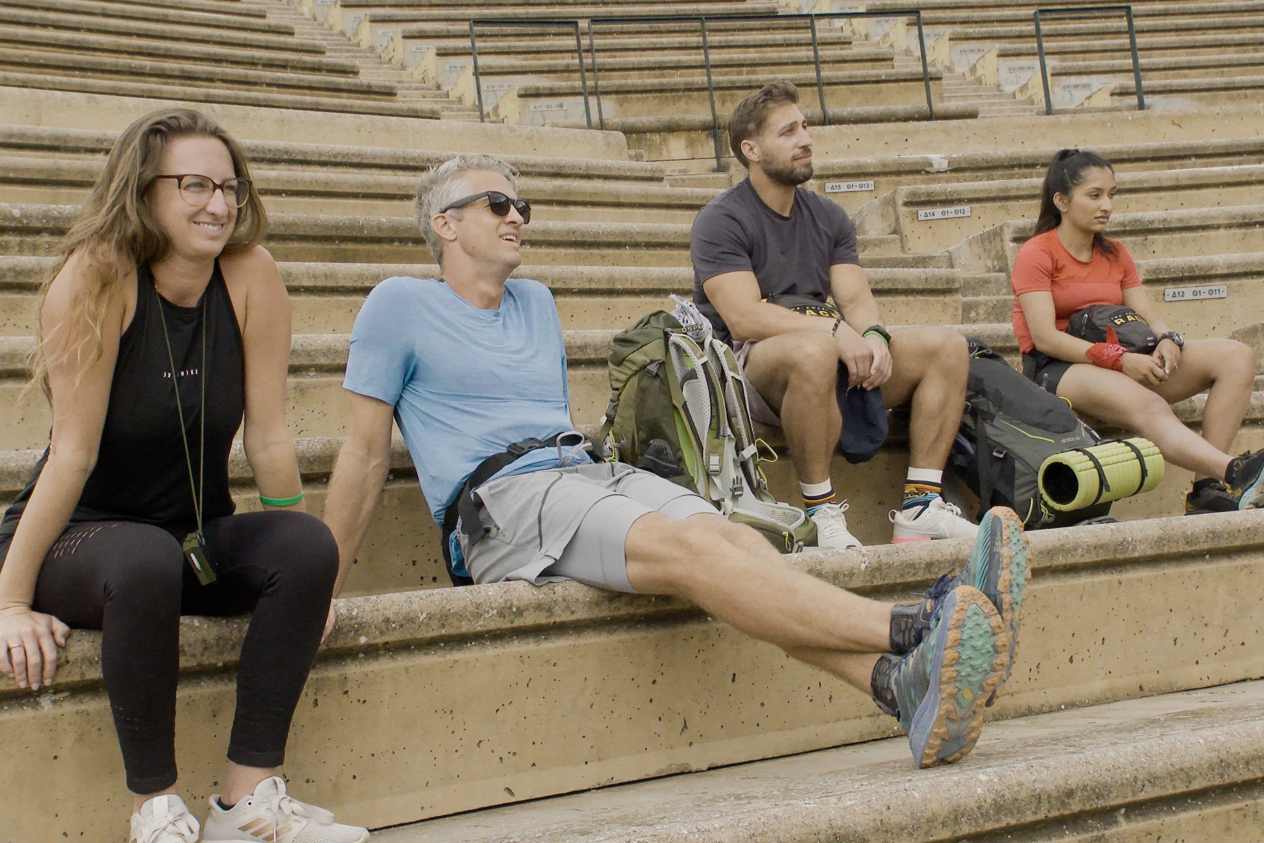 'The Amazing Race' Season 33 contestants Raquel Moore, Penn Holderness, Ryan Ferguson, and Natalia Kumar sit on steps and await the spoilers of who can finish a Roadblock. Raquel wears a black tank top and black leggings. Penn wears a light blue shirt and gray shorts. Ryan wears a dark gray shirt and light gray shorts. Natalia wears a red shirt and black shorts.