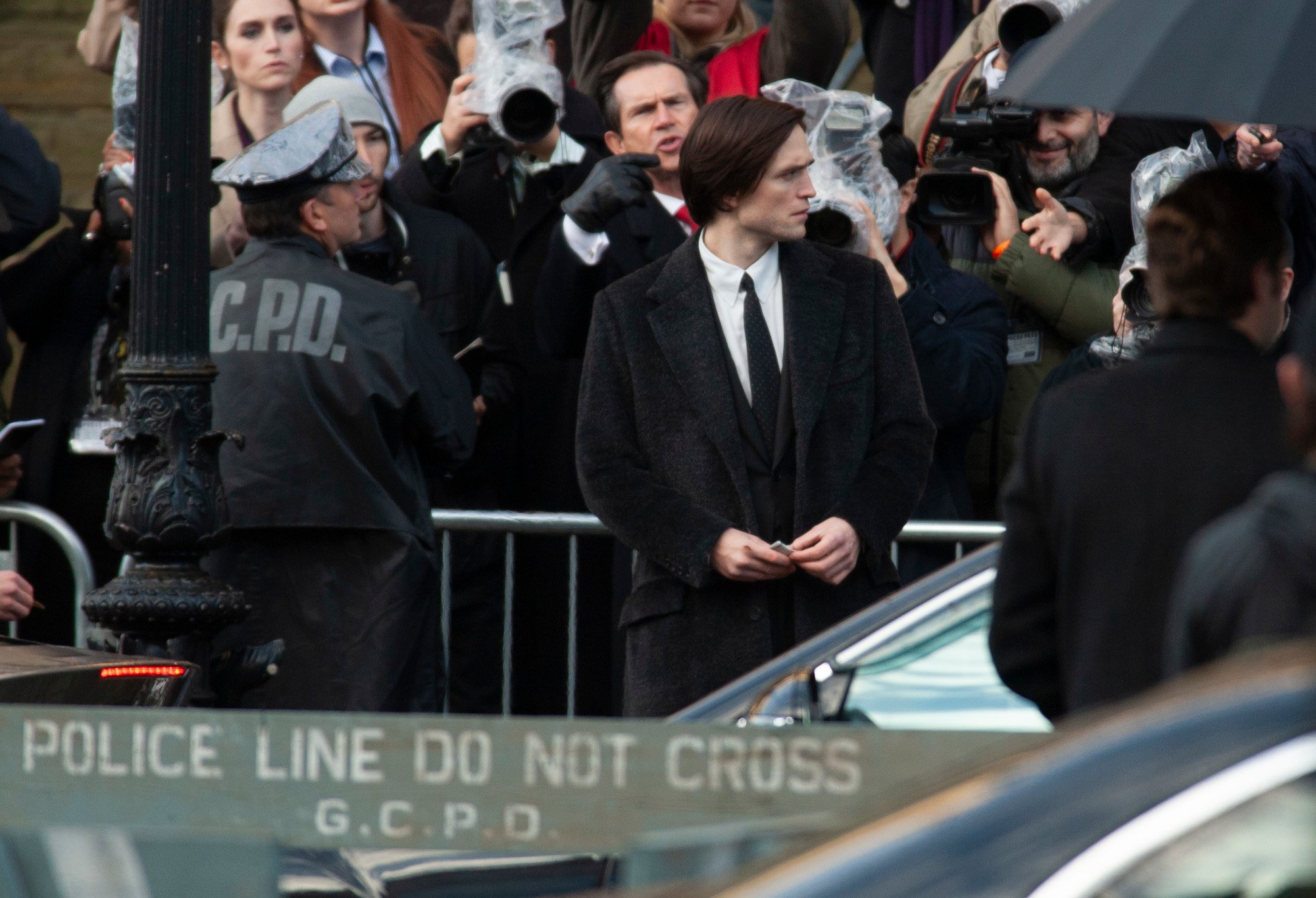 Robert Pattinson on the set of 'The Batman.' He's dressed as Bruce Wayne and surrounded by GCPD offfers and equipment.