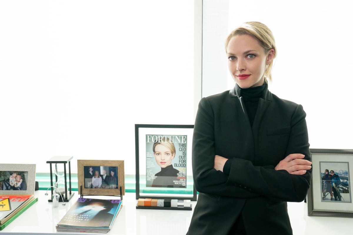 Amanda Seyfried as Elizabeth Holmes in The Dropout Episode 5. Elizabeth stands in front of a framed magazine cover of herself.