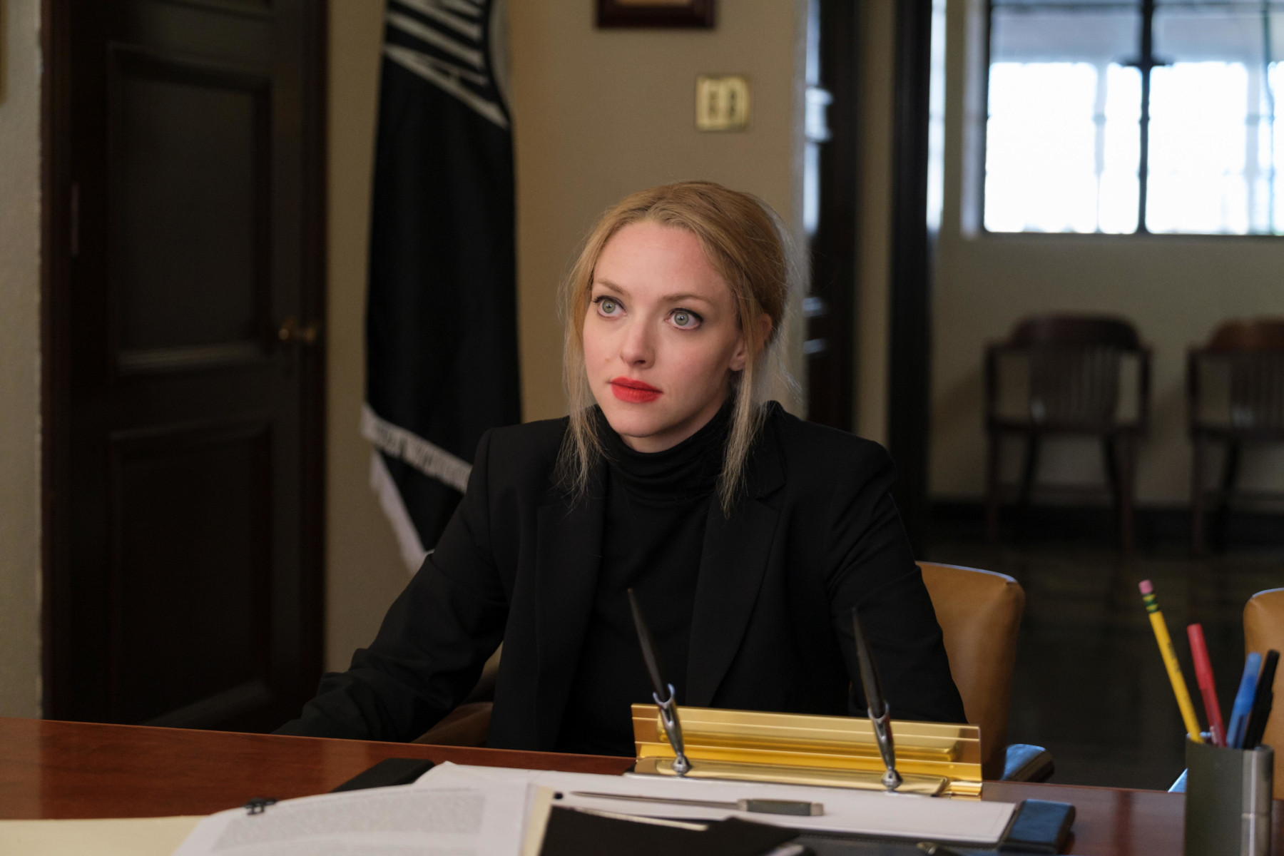 Amanda Seyfried in 'The Dropout' Episode 4 on Hulu. She's wearing a black shirt, red lipstick, and her hair is pulled back. She's sitting in front of a desk.
