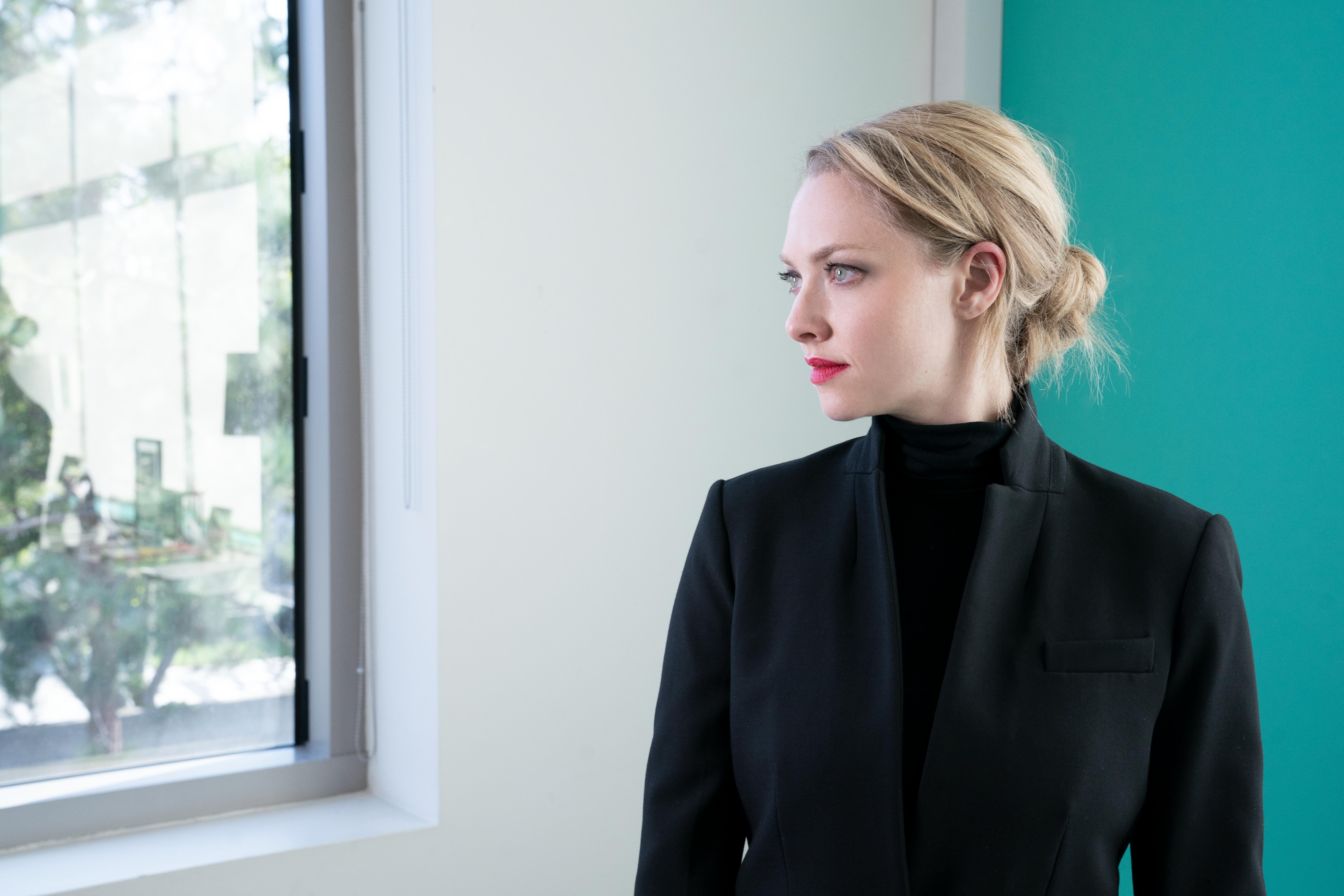 Amanda Seyfried as Elizabeth Holmes in 'The Dropout' Episode 5. She's wearing a black turtle neck and black blazer and standing in front of white and green walls.