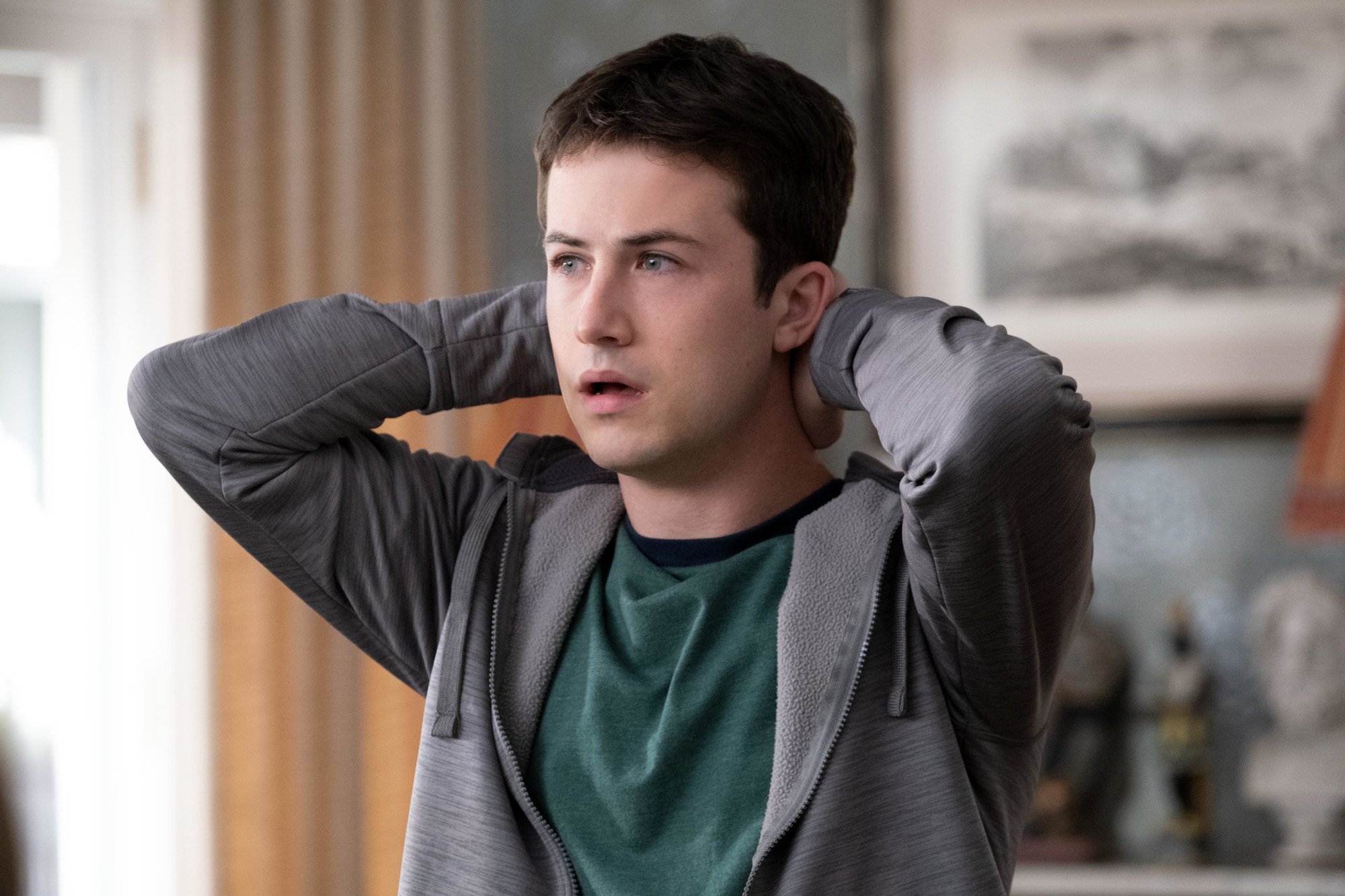 Dylan Minnette as Tyler Shultz in 'The Dropout' Episode 7 on Hulu. He's wearing a green shirt and grey hooded sweatshirt. He looks stressed and his arms are up and behind his head.