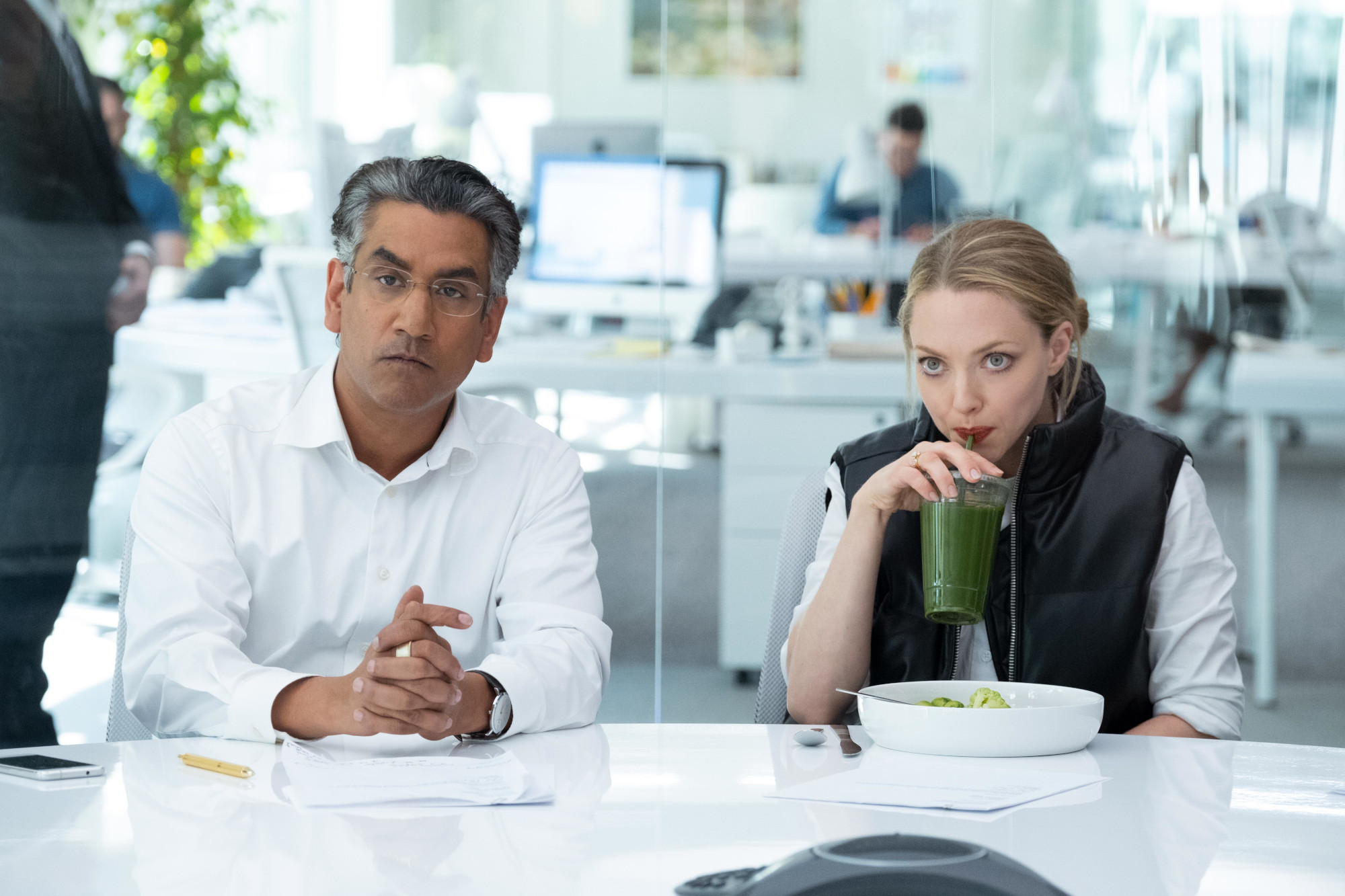 Naveen Andrews and Amanda Seyfried as Sunny Balwani and Elizabeth Holmes in 'The Dropout' Episode 7. They're sitting at a white table, and Seyfried's character is drinking green juice.