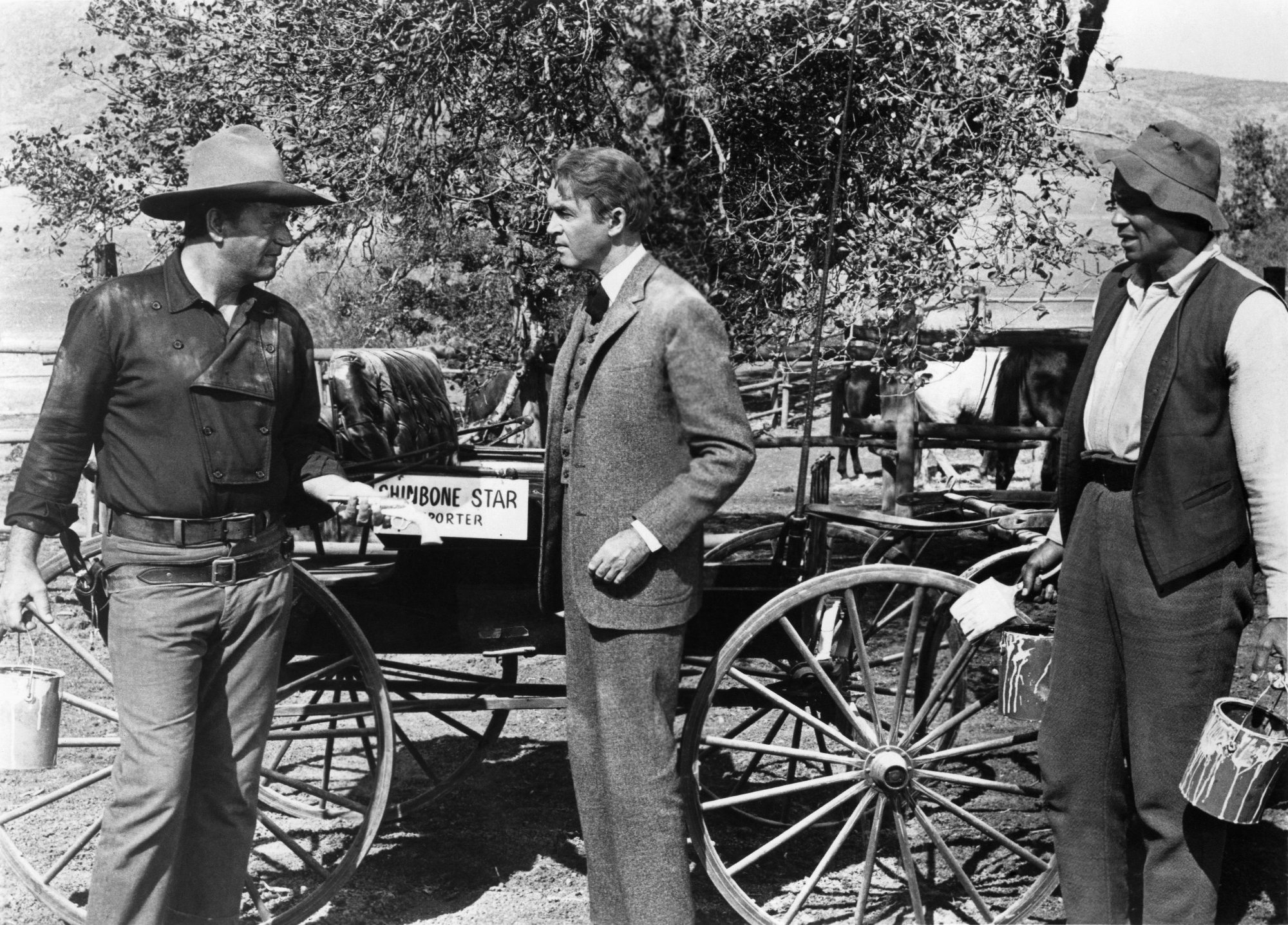 'The Man Who Shot Liberty Valance' John Wayne as Tom Doniphon, James Stewart as Ransom Stoddard, and Woody Strode as Pompey standing in front of a wagon looking at each other in Western costumes