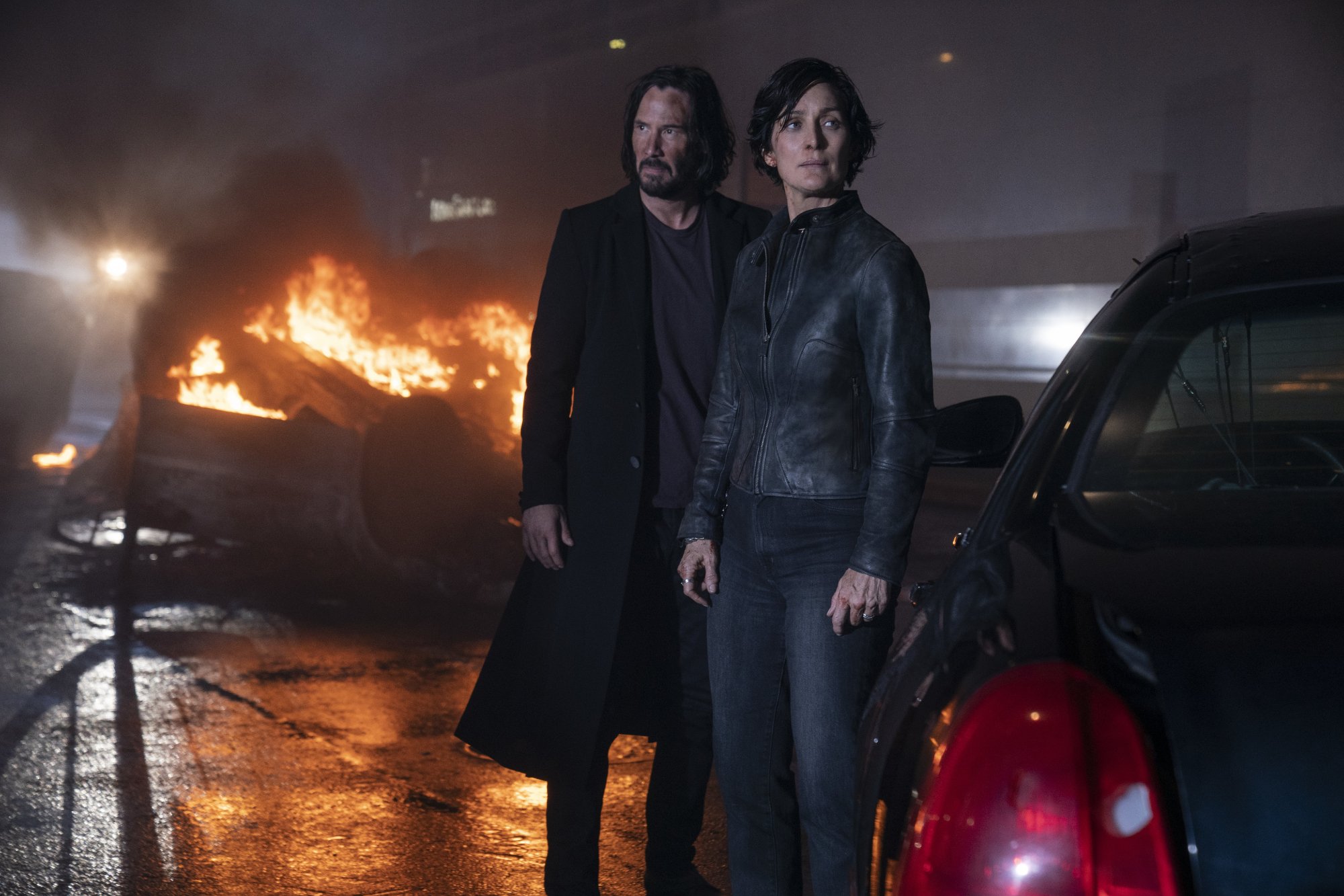 'The Matrix Resurrections' Keanu Reeves as Neo and Carrie-Anne Moss as Trinity after dive bomb climax looking around with a car on fire in the background