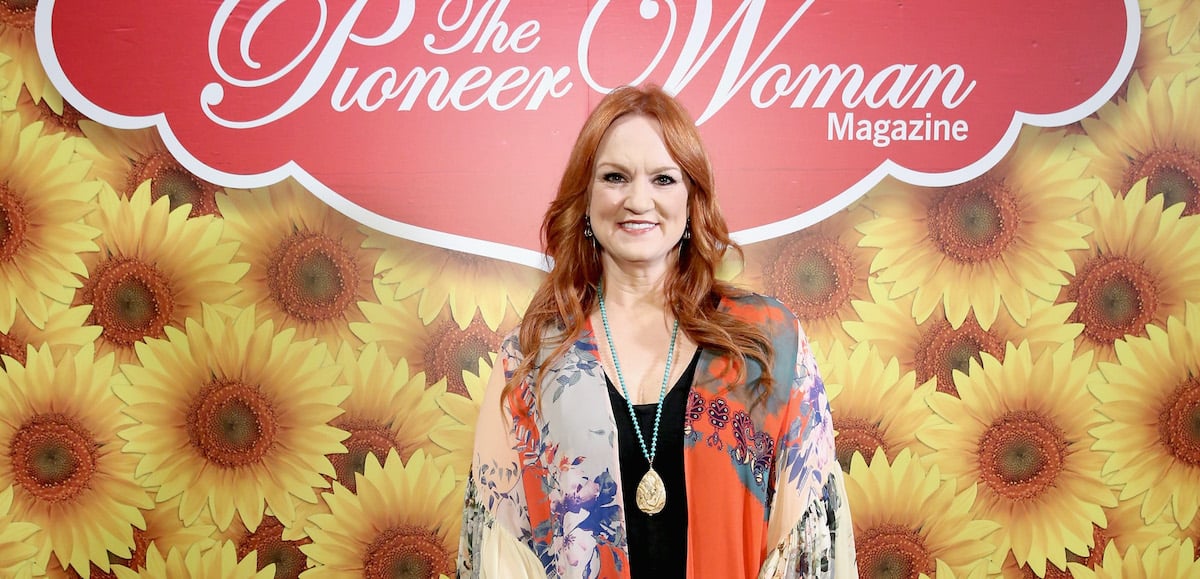 Pioneer Woman Ree Drummond smiles wearing a black shirt and multicolor sweater in front a Pioneer Woman Magazine sign